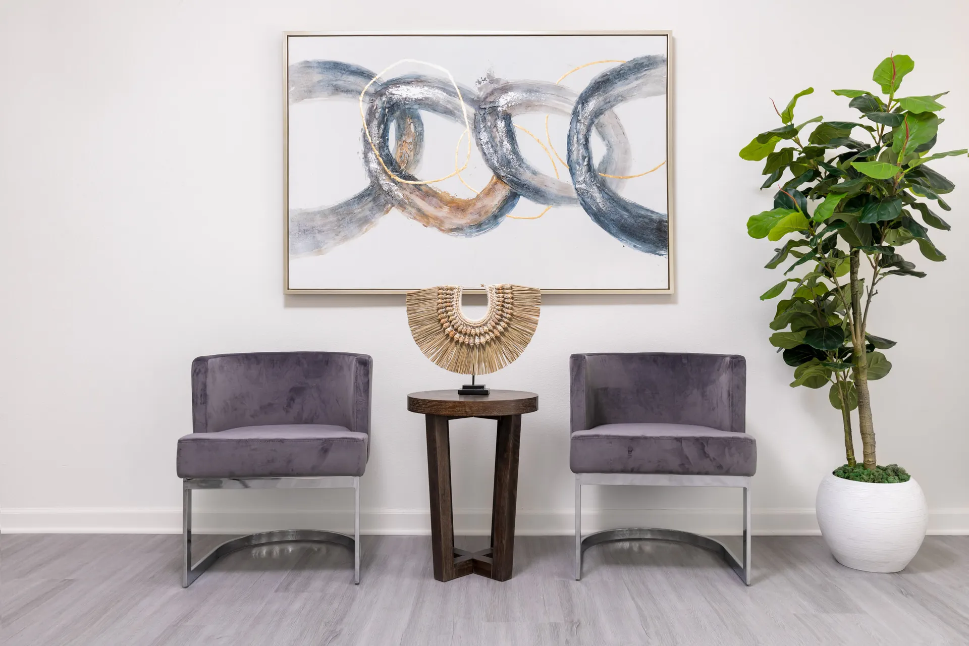 Two gray velvet chairs flank a small round table displaying a decorative accent under an abstract painting, with a tall indoor plant to the right.