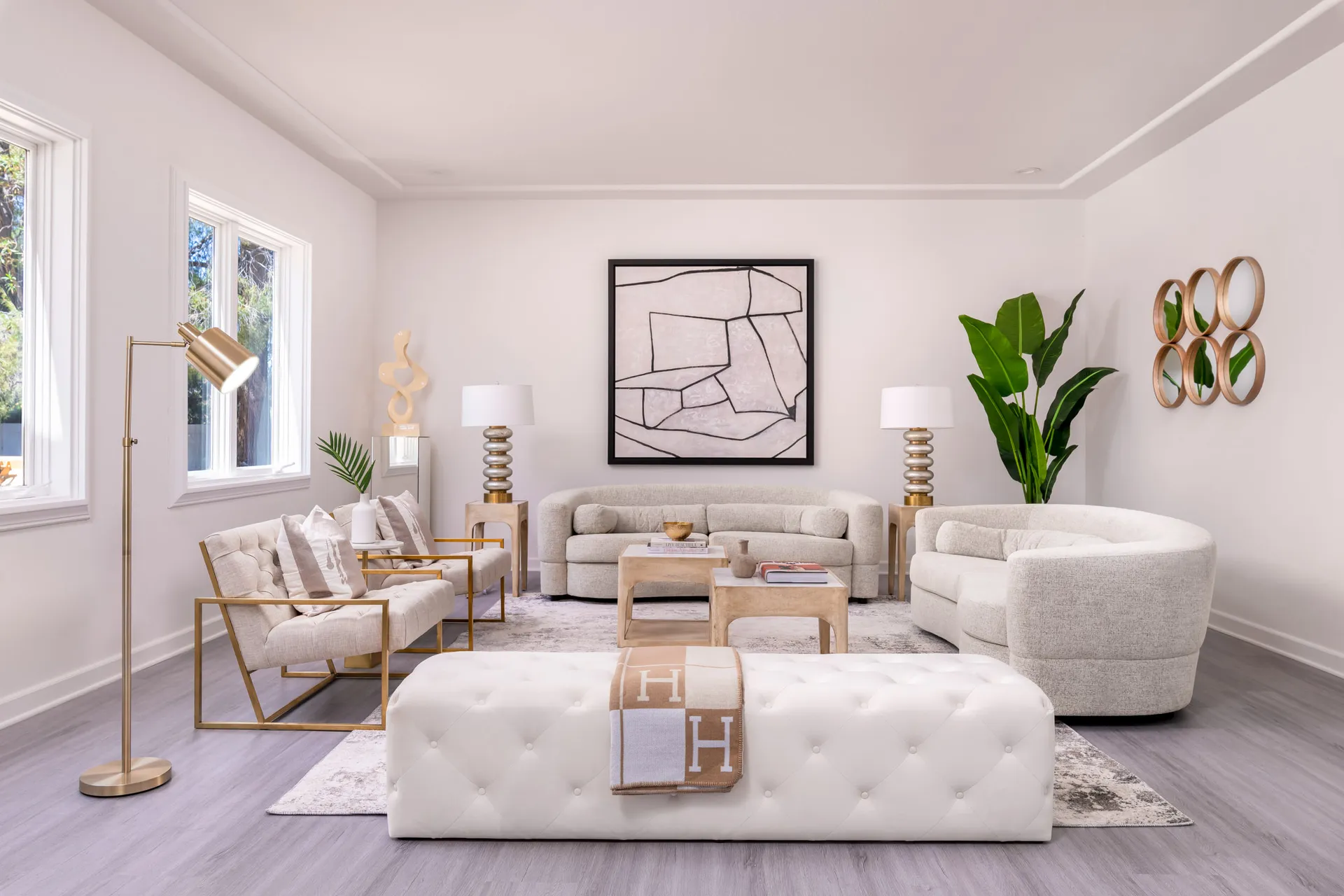 A light-filled living room with two curved sofas, a pair of gold-framed chairs, a tufted white bench, and a large abstract painting.