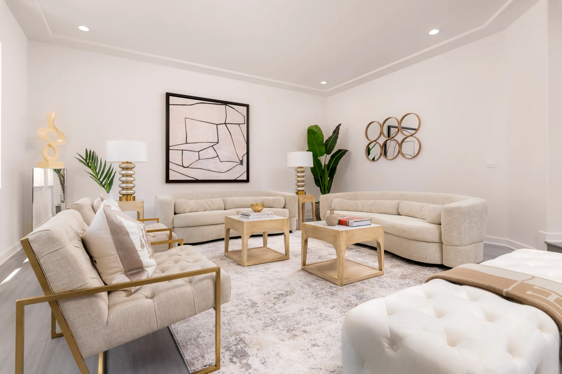 A neutral living area with curved beige sofas, tufted chairs featuring gold frames, wooden coffee tables, and a large abstract painting.