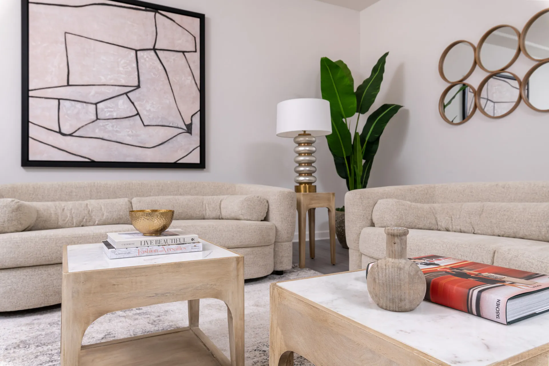 Two curved beige sofas facing marble-topped wooden tables, with an abstract wall art piece and a tall green plant in the background.