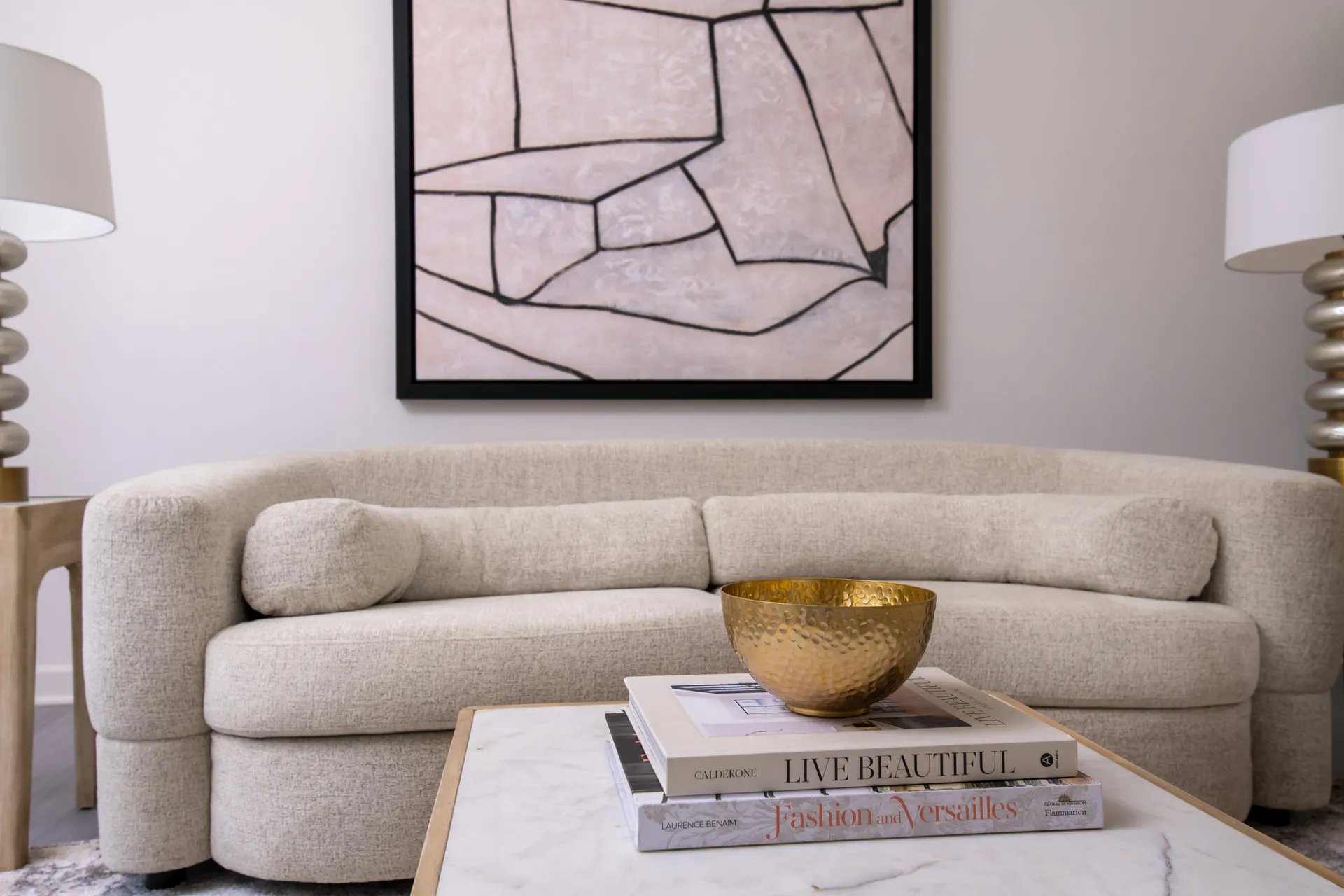 A curved beige sofa with round accent pillows in front of an abstract painting. A marble-topped table holds a stack of books and a gold bowl.