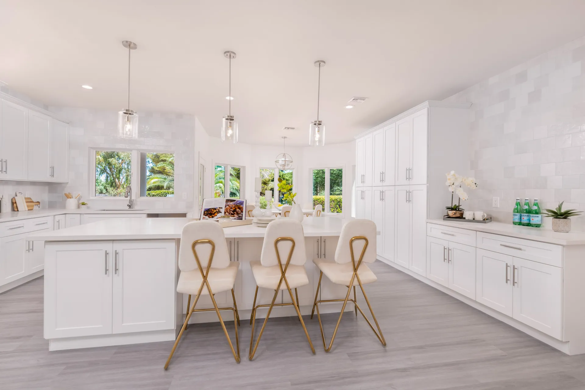 A spacious white kitchen with a large island, gold barstools, and ample cabinetry under bright windows.
