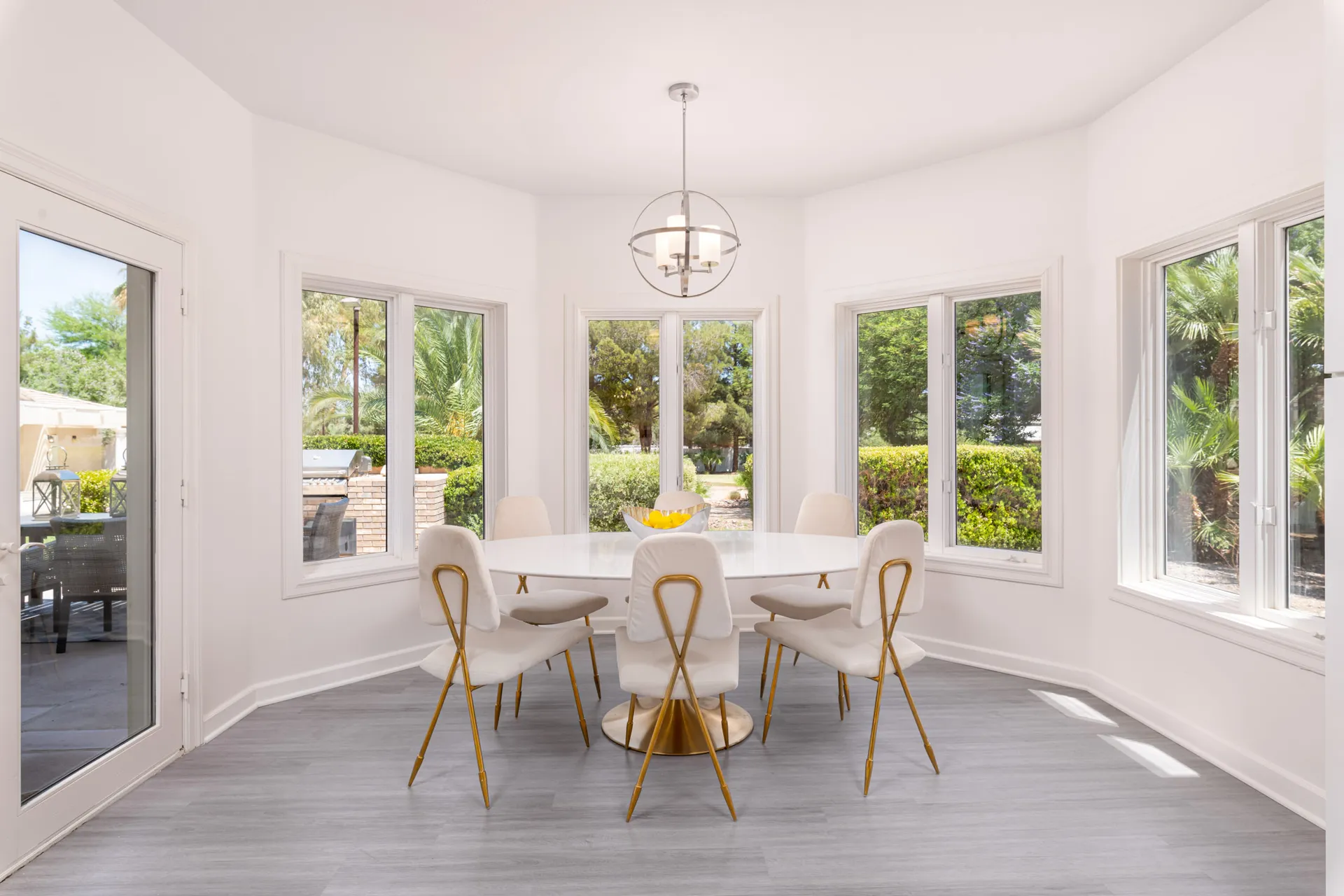 A round white dining table with gold-accent chairs, surrounded by windows overlooking lush greenery.