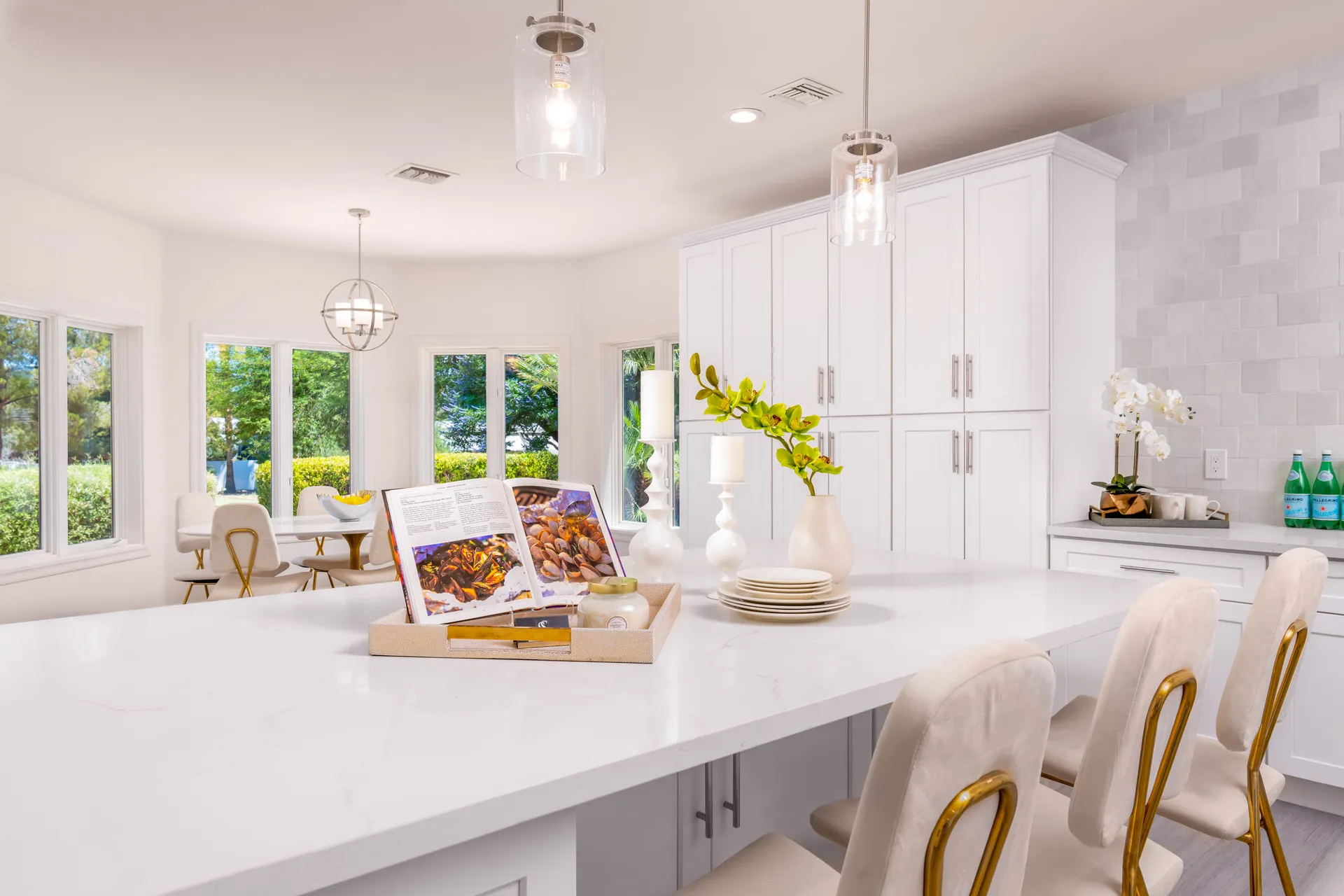 A bright kitchen island with white countertops, gold-accent chairs, and a tray holding an open cookbook, candles, and greenery.