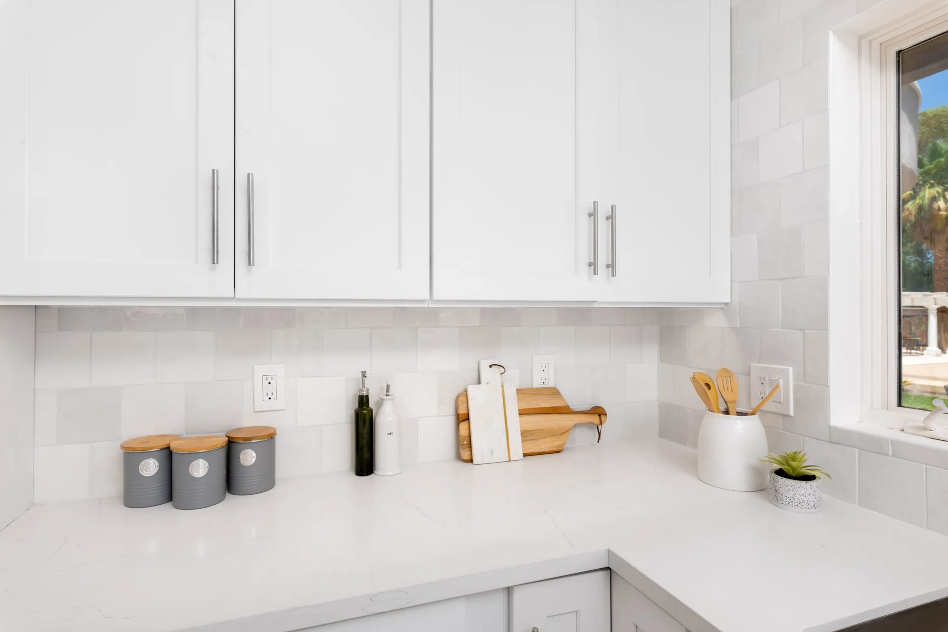 A white kitchen counter with canisters, oil bottles, and a marble cutting board under white cabinets.