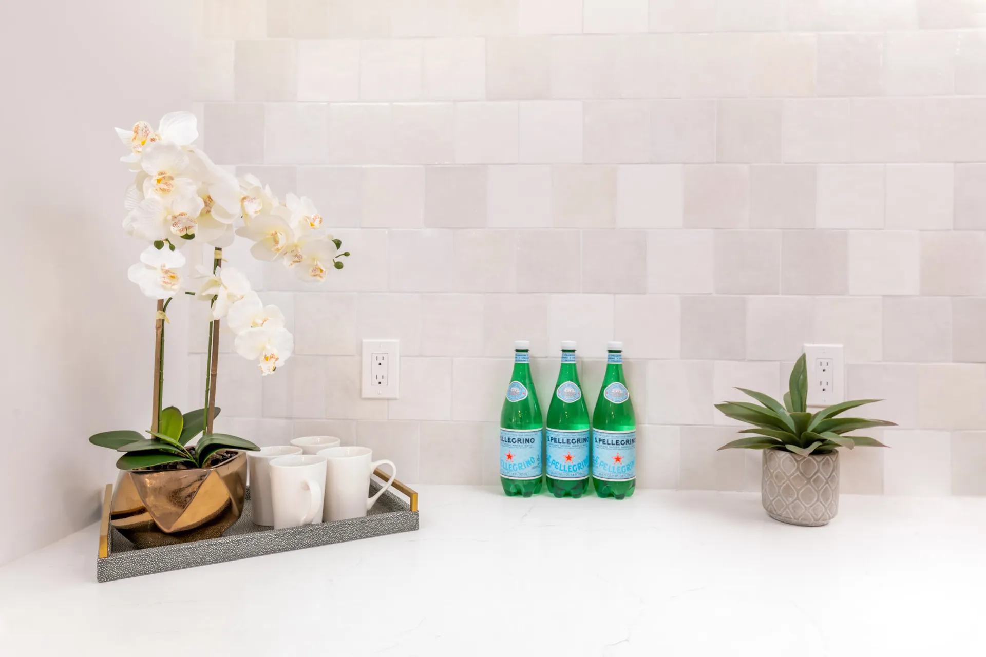 A gold potted orchid, white mugs on a tray, and sparkling water bottles on a white countertop.