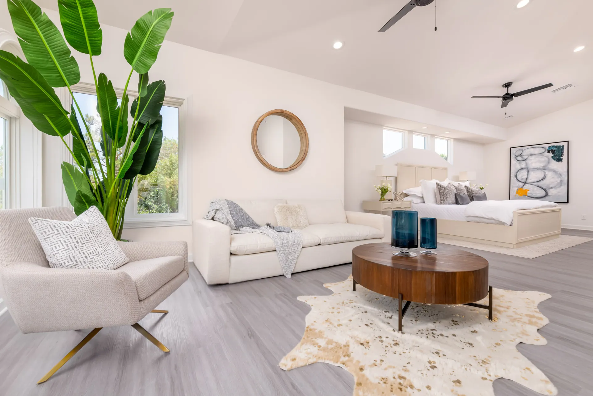 A bright bedroom-lounge area with a neutral sofa, a round wooden coffee table, a tall plant, and a bed in the background.