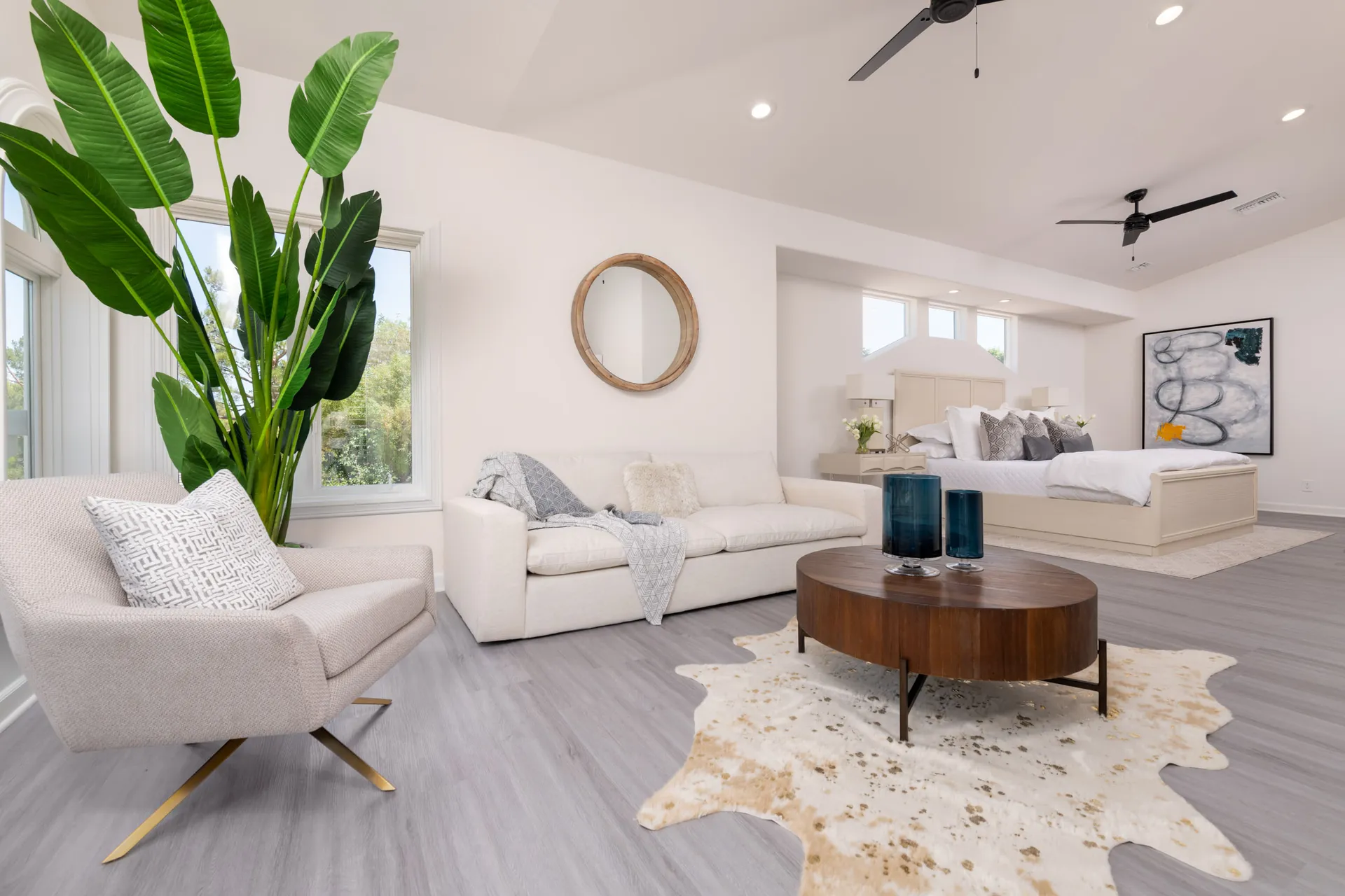 A spacious bedroom with a neutral-toned bed, a cozy sofa, a round wooden coffee table, and a tall green plant near large windows.