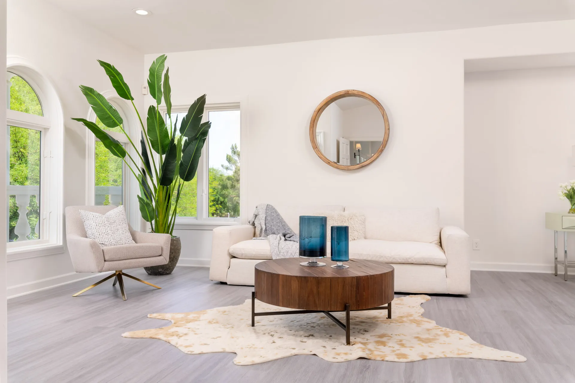A cozy seating area with a beige sofa, a round wooden coffee table, a tall green plant, and large windows allowing ample natural light.