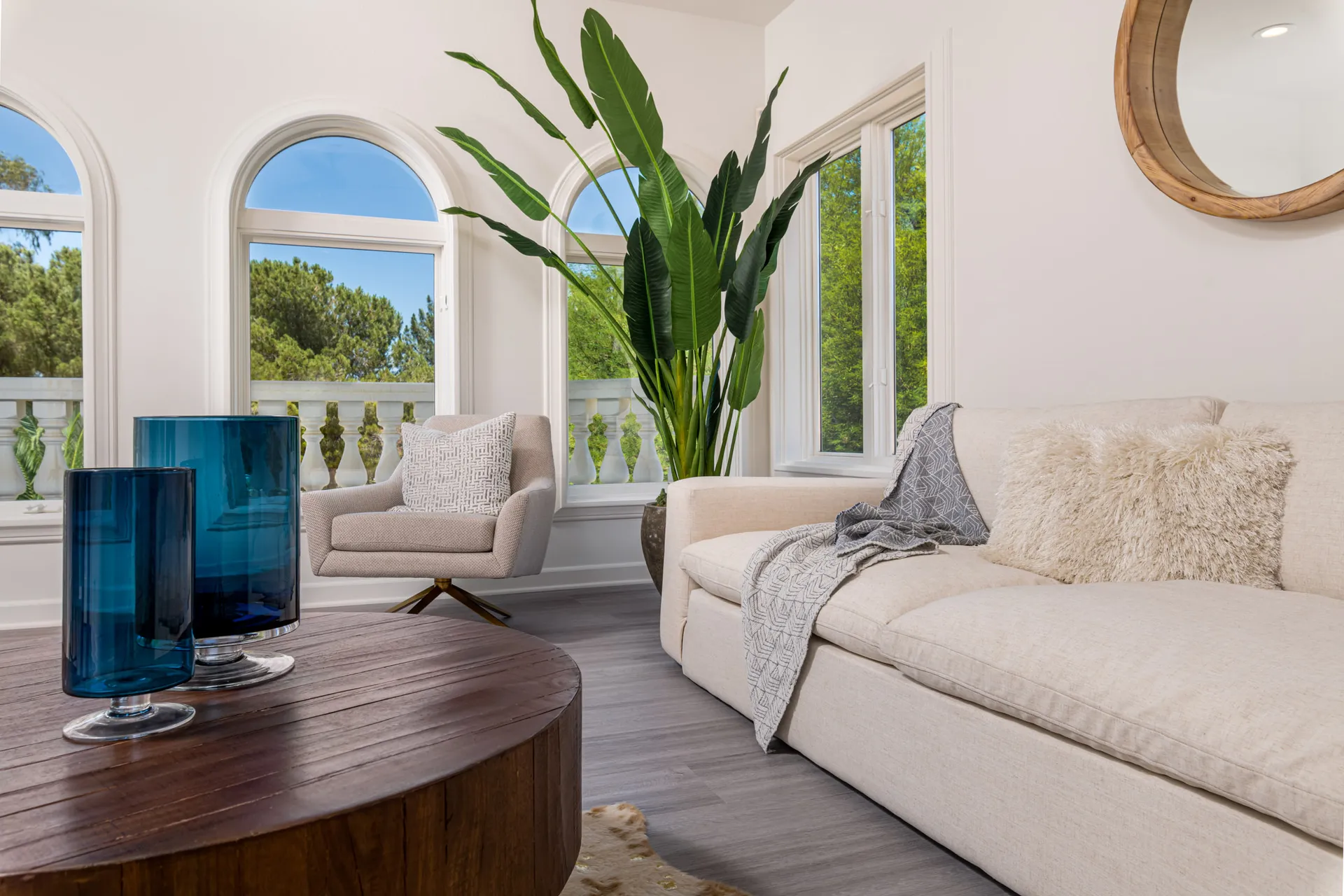A cozy lounge area featuring a beige sofa, a round wooden table with blue glass vases, and large windows framing lush greenery.