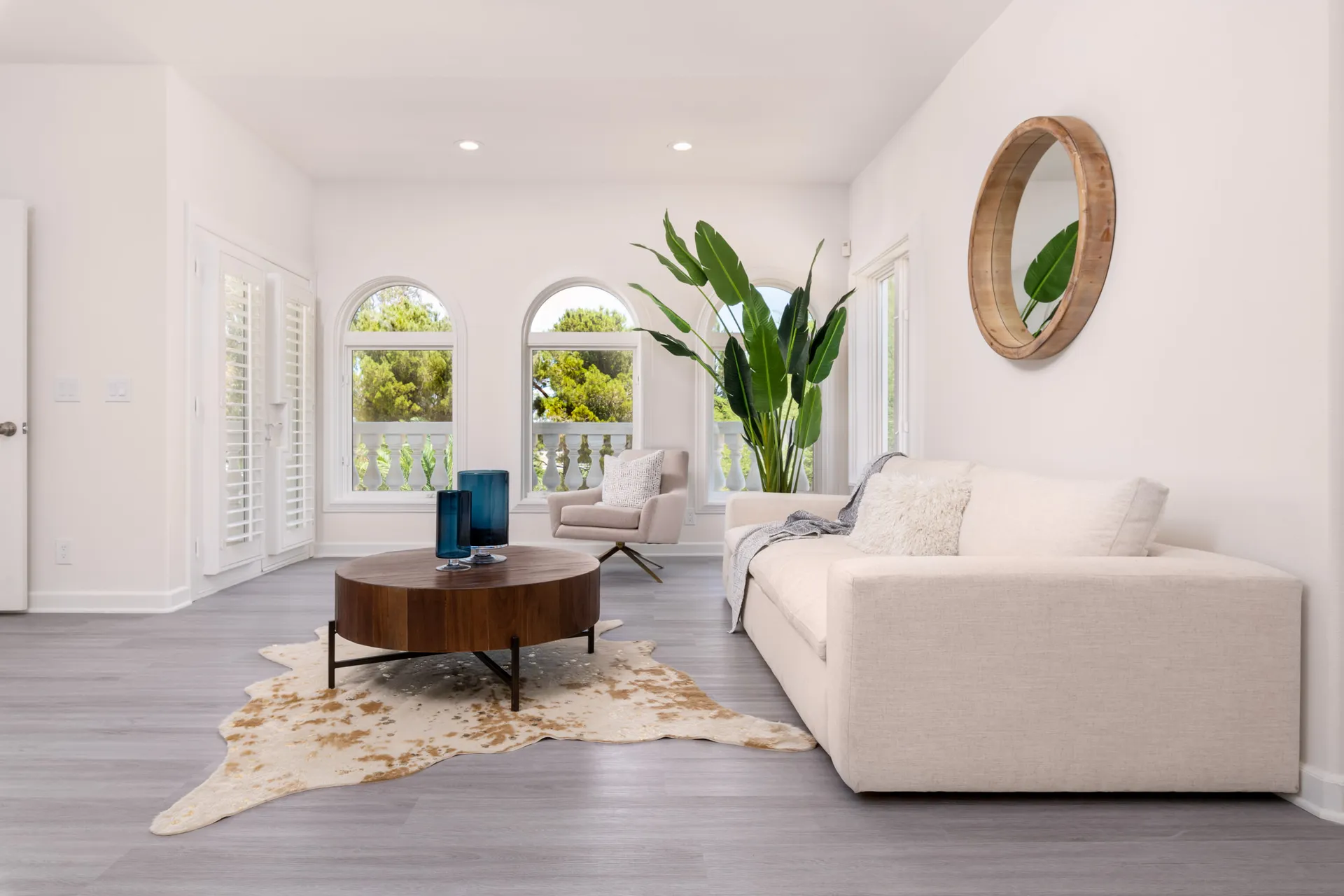 A cozy seating nook with a beige sofa, round wooden coffee table, arched windows, and a tall green plant.