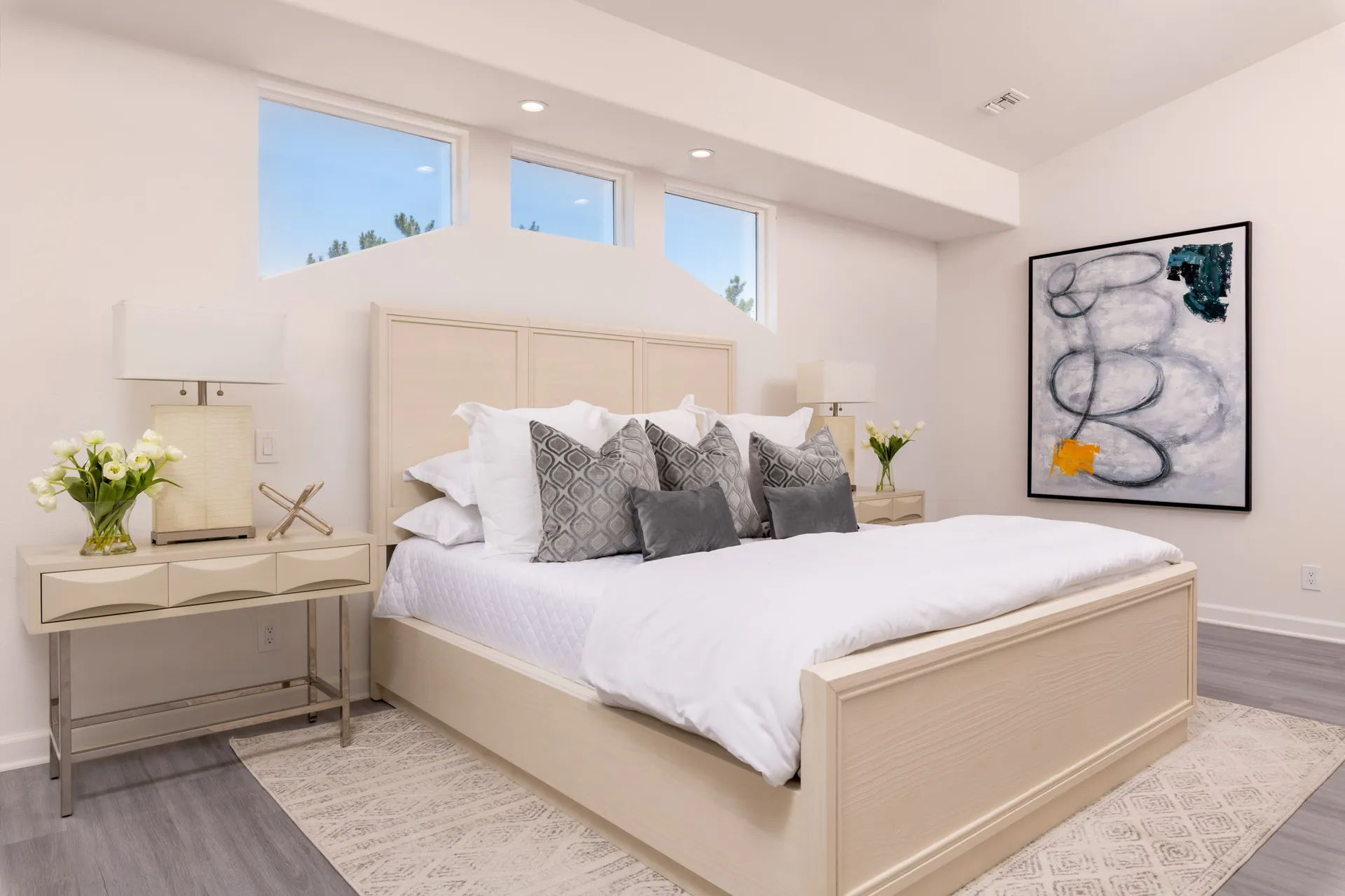 A neutral bed with crisp white linens, layered gray pillows, and an abstract painting on the wall.