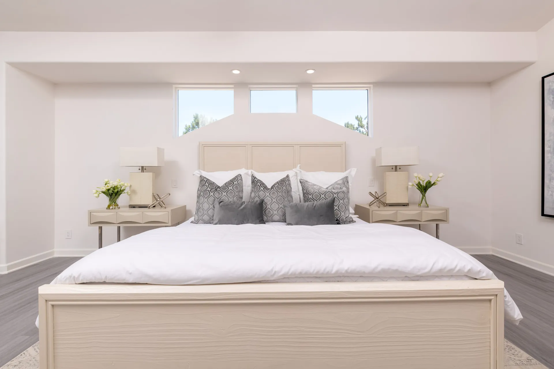 A light-toned bed with crisp white linens, gray throw pillows, and matching beige nightstands under high windows.
