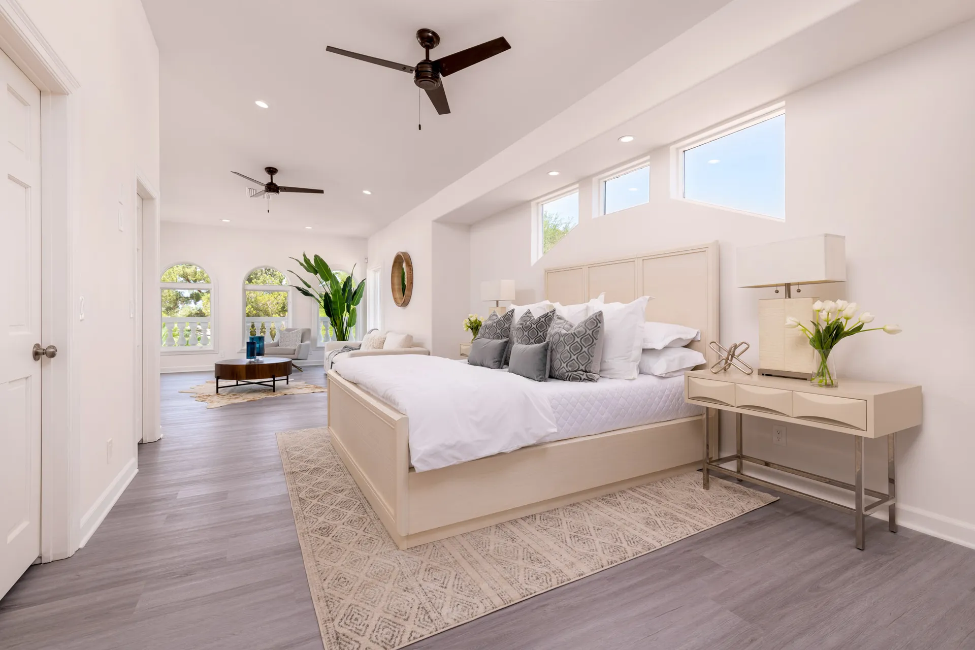 A bright bedroom with a light wood bed, white linens, ceiling fans, and a separate seating area by large windows.