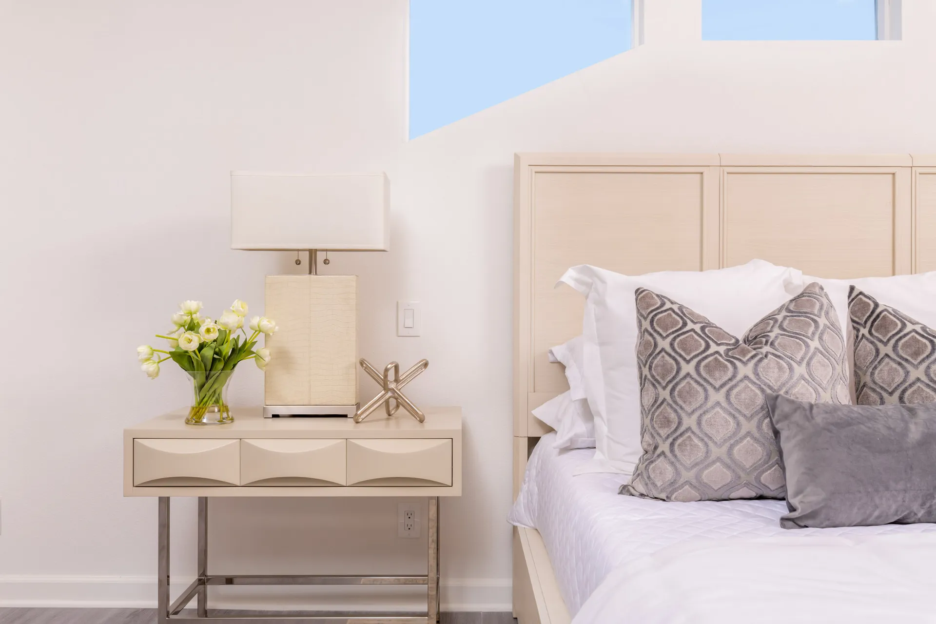 A light-colored bed with plush pillows, a beige nightstand, and a vase of tulips.