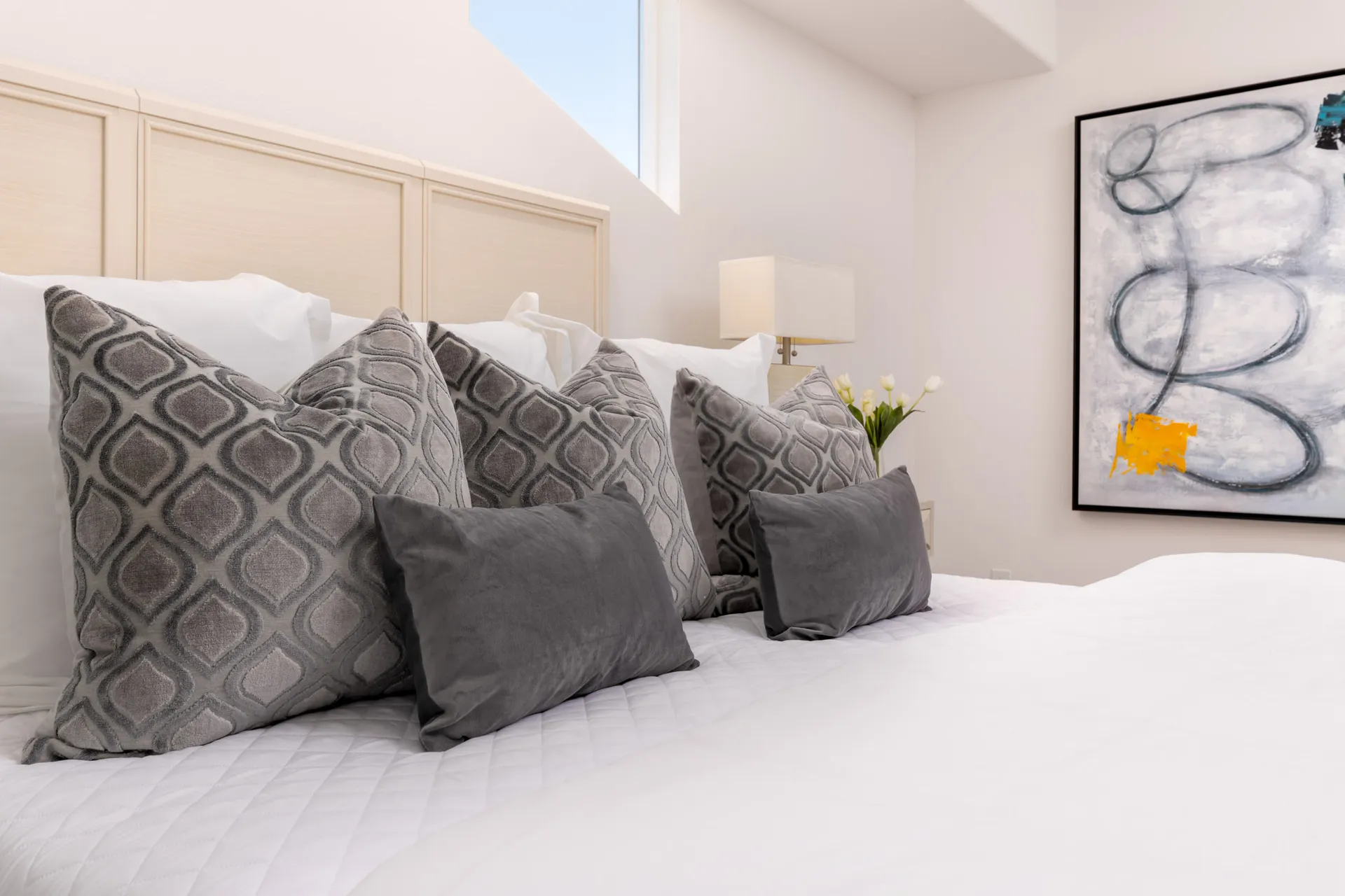 A bed with white linens and layered gray accent pillows near a large abstract painting.