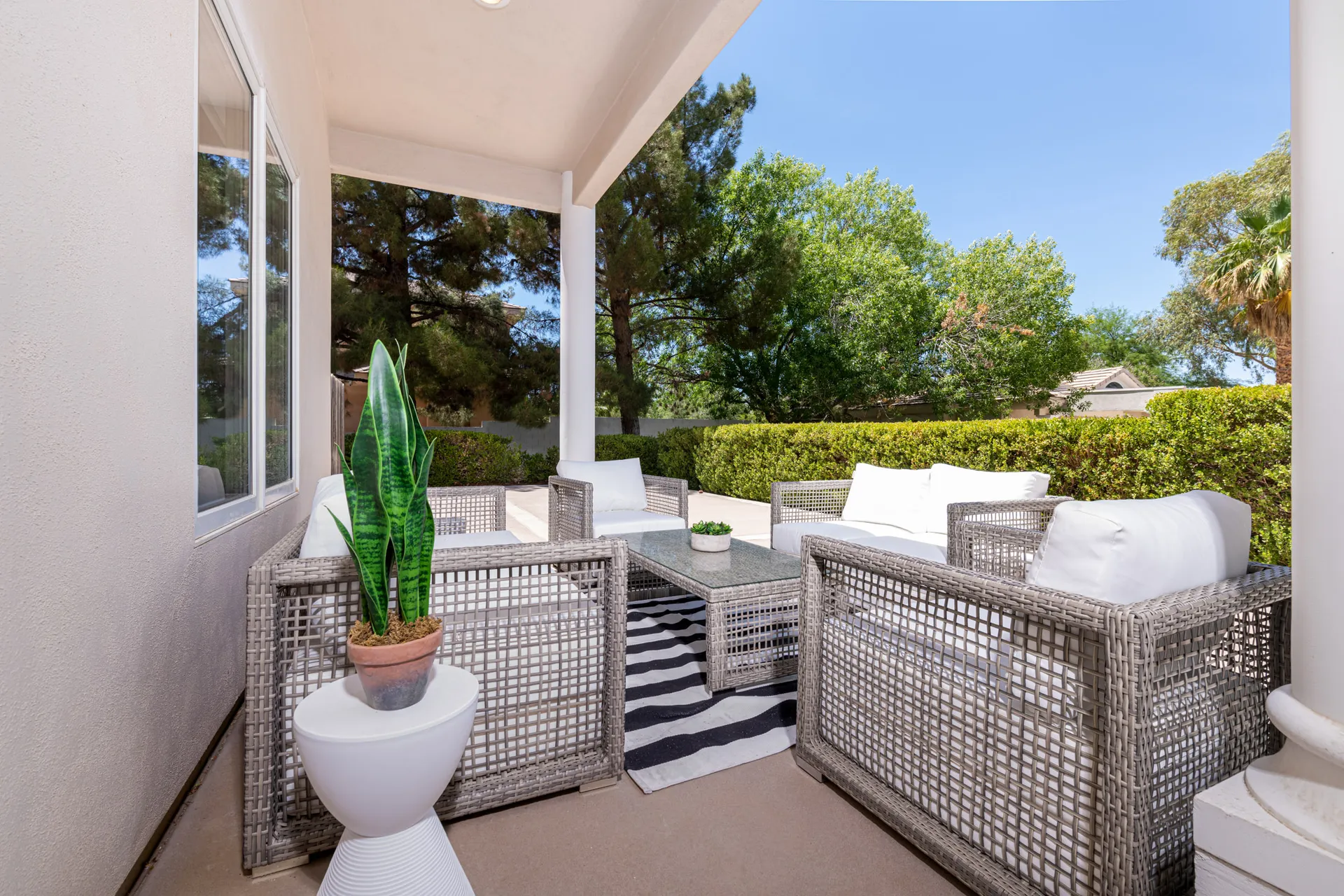 A covered patio with wicker chairs, white cushions, a striped rug, and a potted plant, surrounded by greenery.
