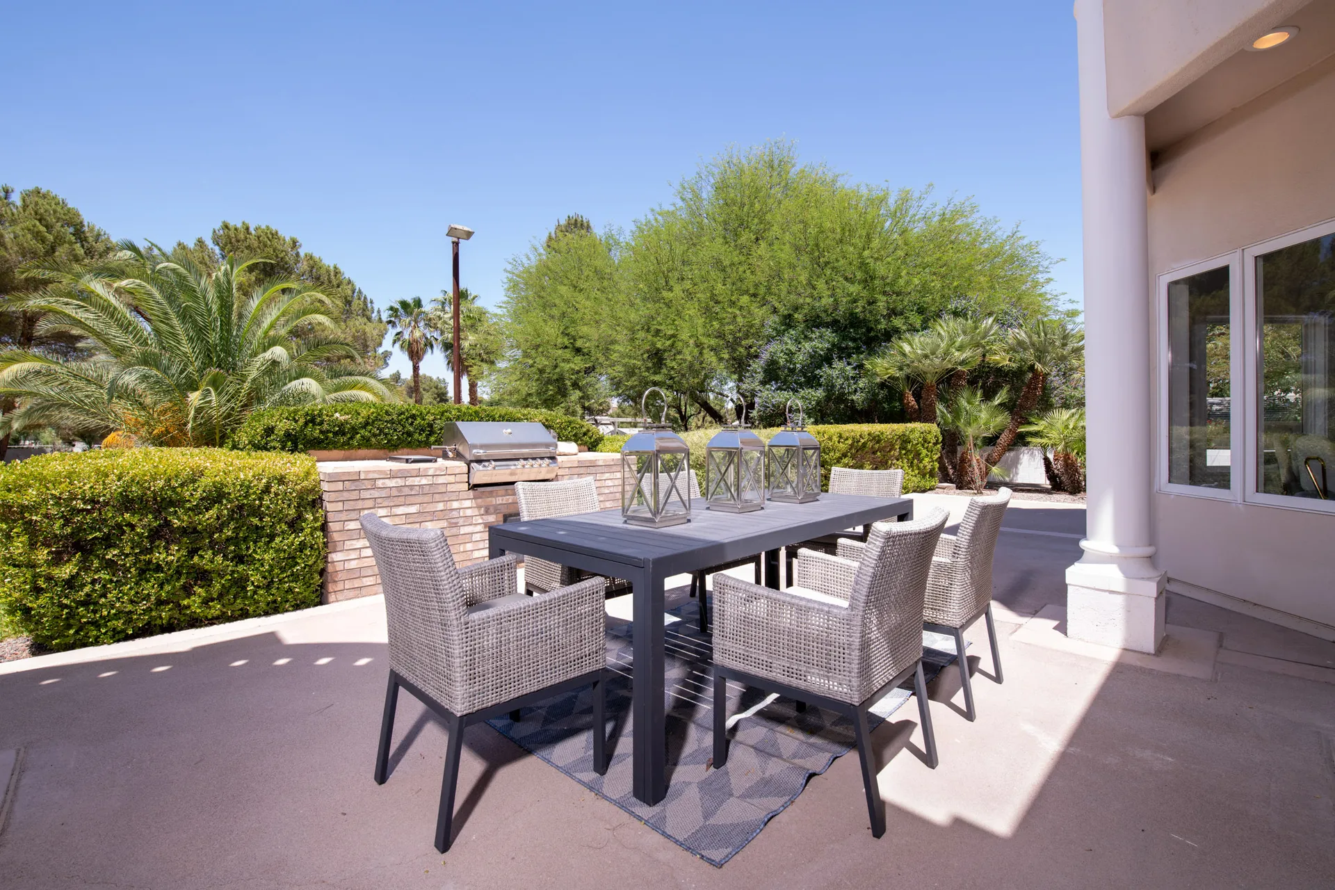 A gray dining table with wicker chairs and lantern centerpieces near a built-in grill on a sunlit patio.