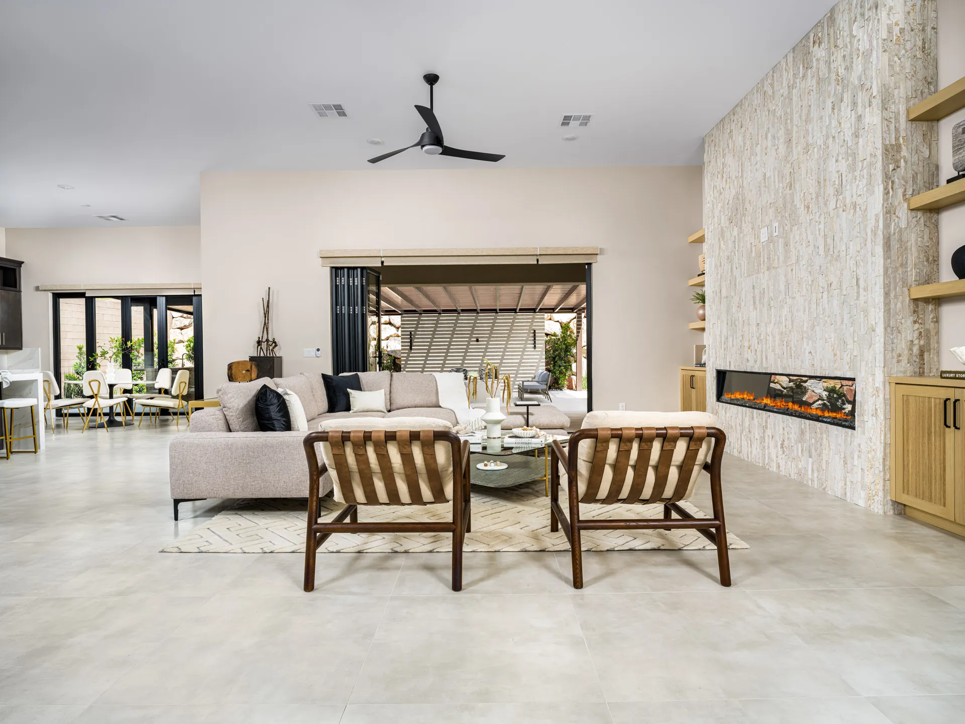 A large open-concept living area with a neutral sectional sofa, stone fireplace, and built-in shelves. The space extends to a dining area with gold-accented seating.
