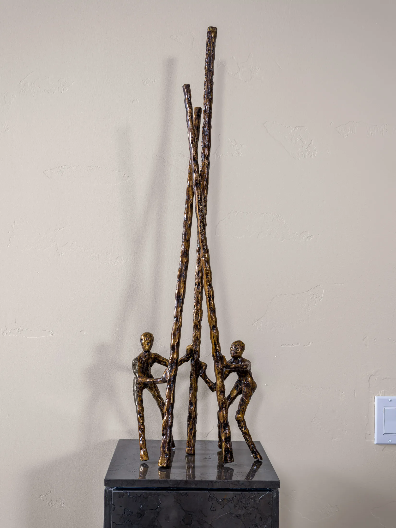 A bronze figurative sculpture showing three people supporting tall, textured rods. The piece rests on a dark pedestal against a neutral wall.