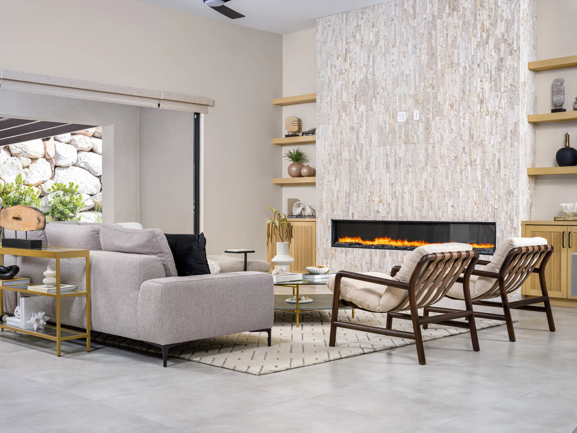A contemporary living area with a large stone fireplace, built-in shelves, neutral seating, and a gold console table. Sliding doors open to an outdoor space.