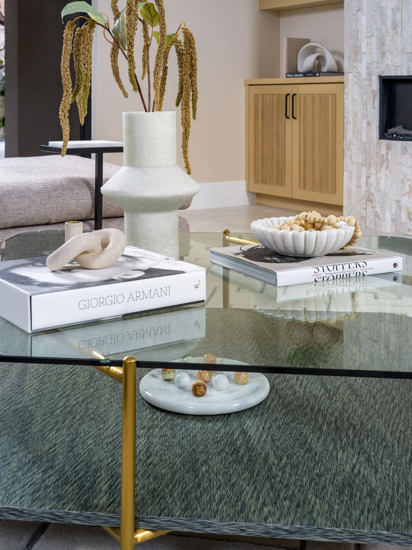 A round glass coffee table with gold legs, featuring a tall cream vase of draping greenery, a looped sculpture, books, and a marble dish of decorative marbles.