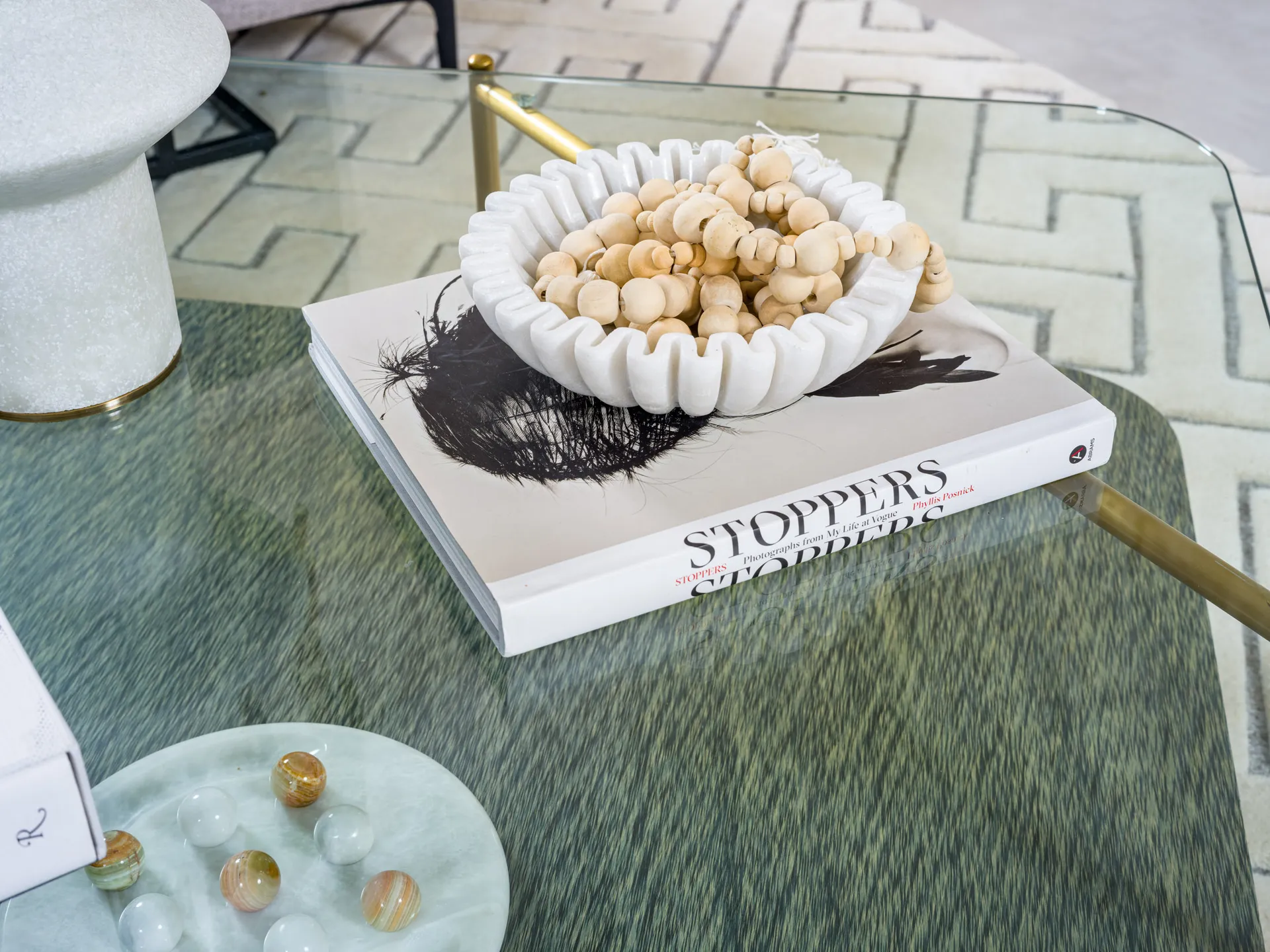 A glass coffee table topped with a hardcover book and a ruffled ceramic bowl filled with wooden beads. The table has gold-toned legs for a touch of elegance.