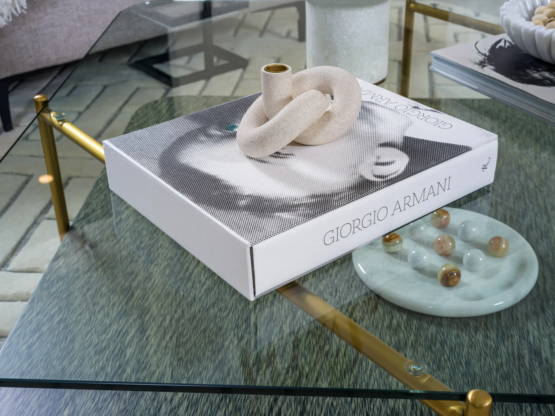 A glass coffee table with a Giorgio Armani book and a looped sculpture on top. A small marble dish with decorative stones is placed nearby.
