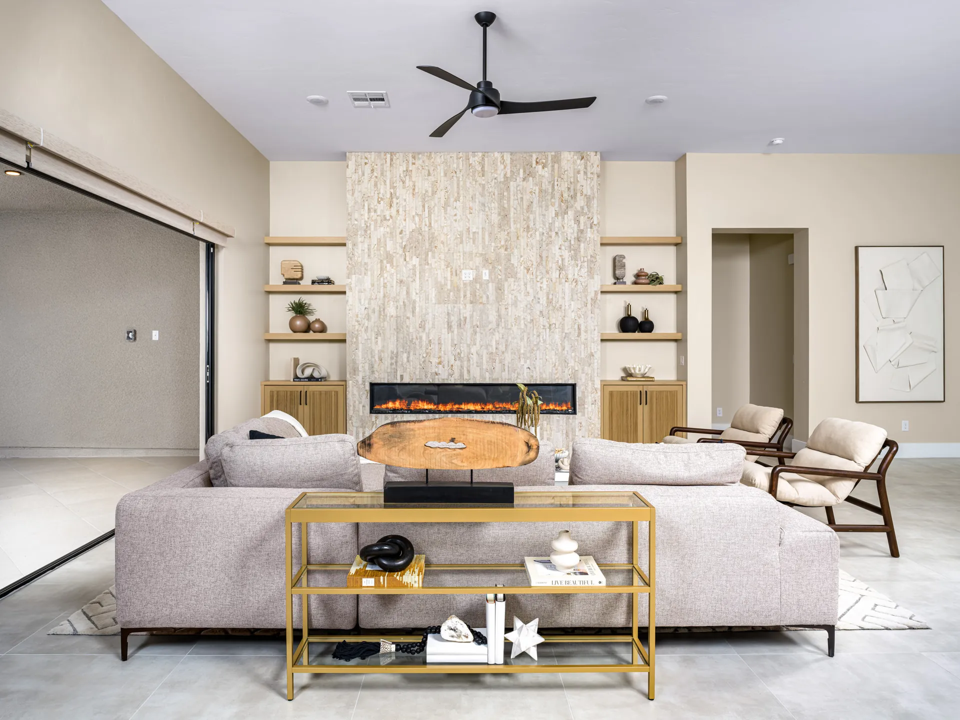 A contemporary living area featuring a stone fireplace wall, built-in shelves, and a gold console table behind a light gray sectional sofa. Large sliding doors open to the outdoors.