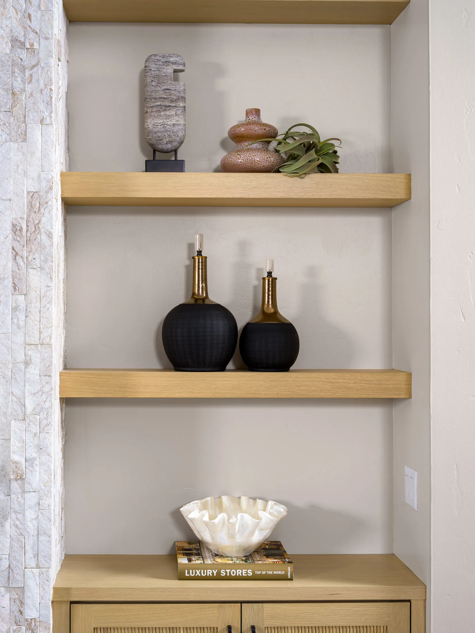 Built-in wooden shelves displaying sculptural pieces, including black-and-gold vases, a stone ornament, and a translucent ruffled bowl on a hardcover book.
