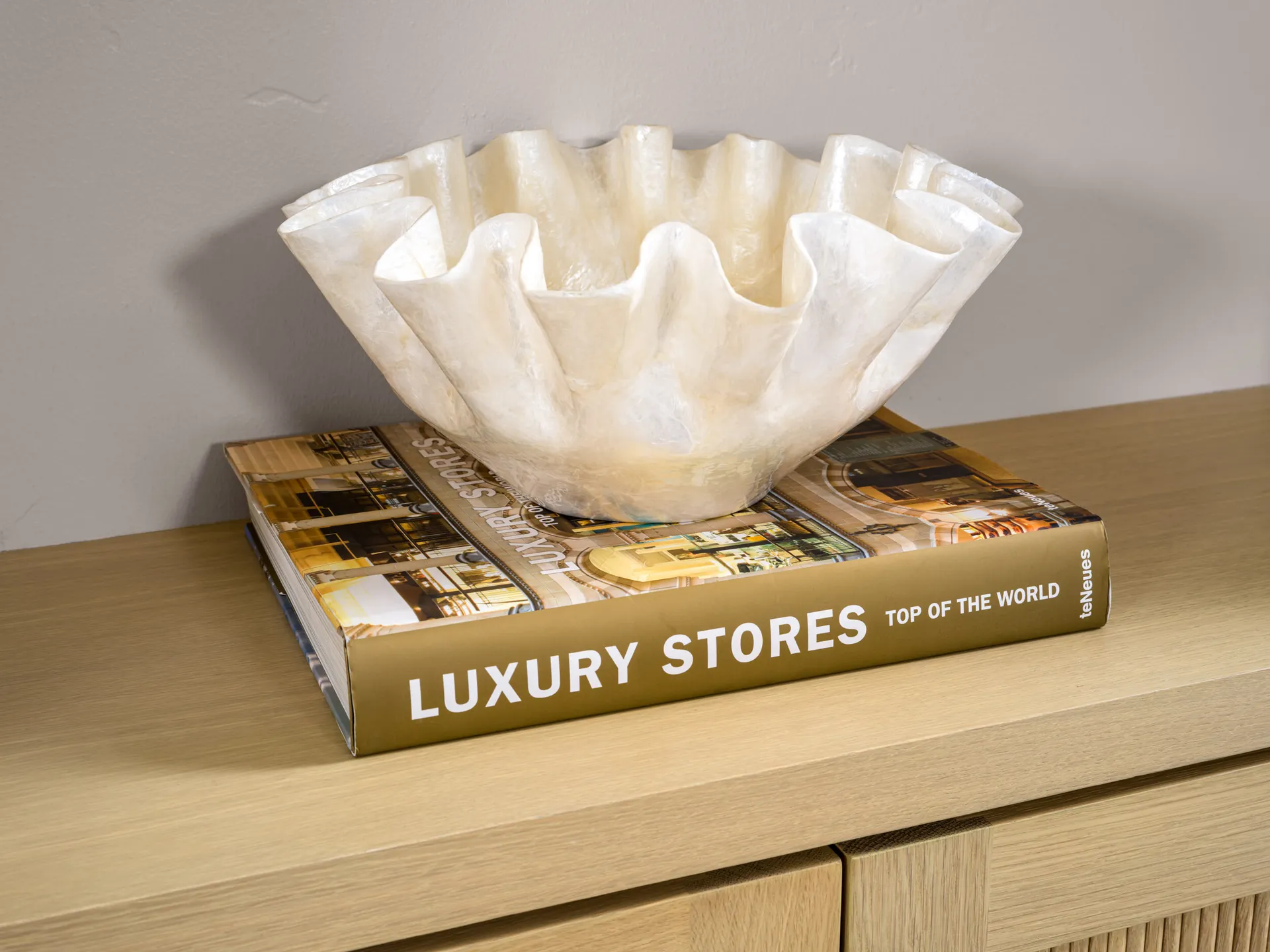 A ruffled, translucent decorative bowl resting on a hardcover book titled ‘Luxury Stores Top of the World,’ placed on a wooden console.