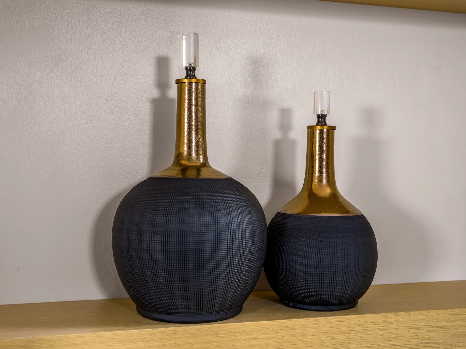 A pair of decorative, bottle-shaped vessels with matte black bases and gold metallic necks. They are displayed on a wooden shelf.