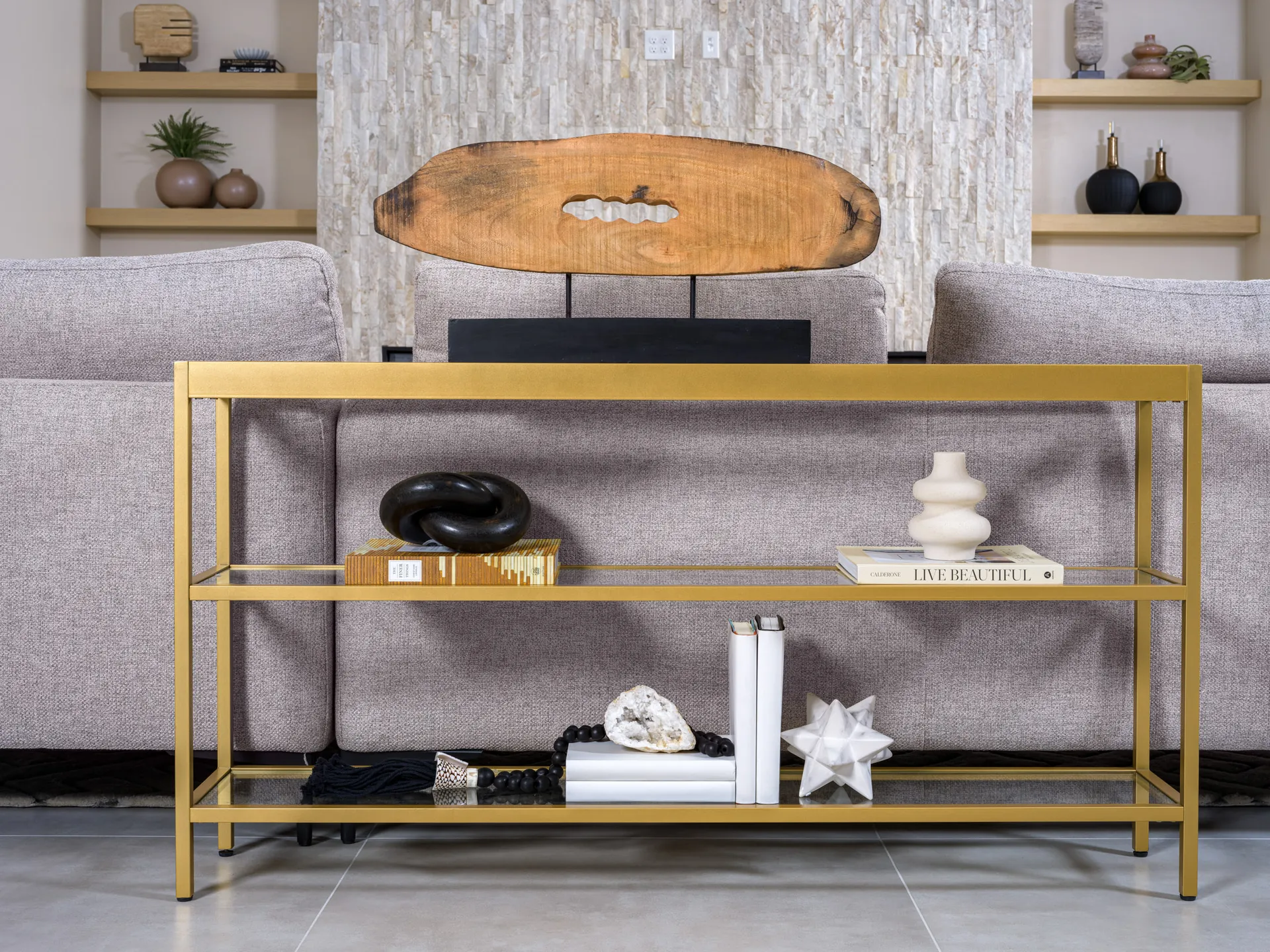 A gold console table behind a gray sofa displaying a wooden sculpture, stacked books, a black knot accent, and a geode alongside other decorative objects.