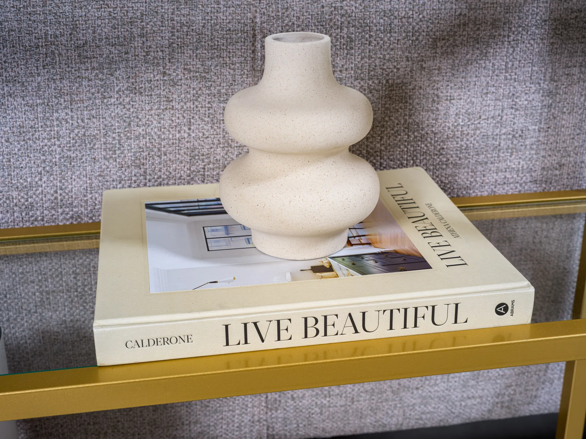A gold-framed shelf displaying a cream-colored sculptural vase resting on a hardcover book.