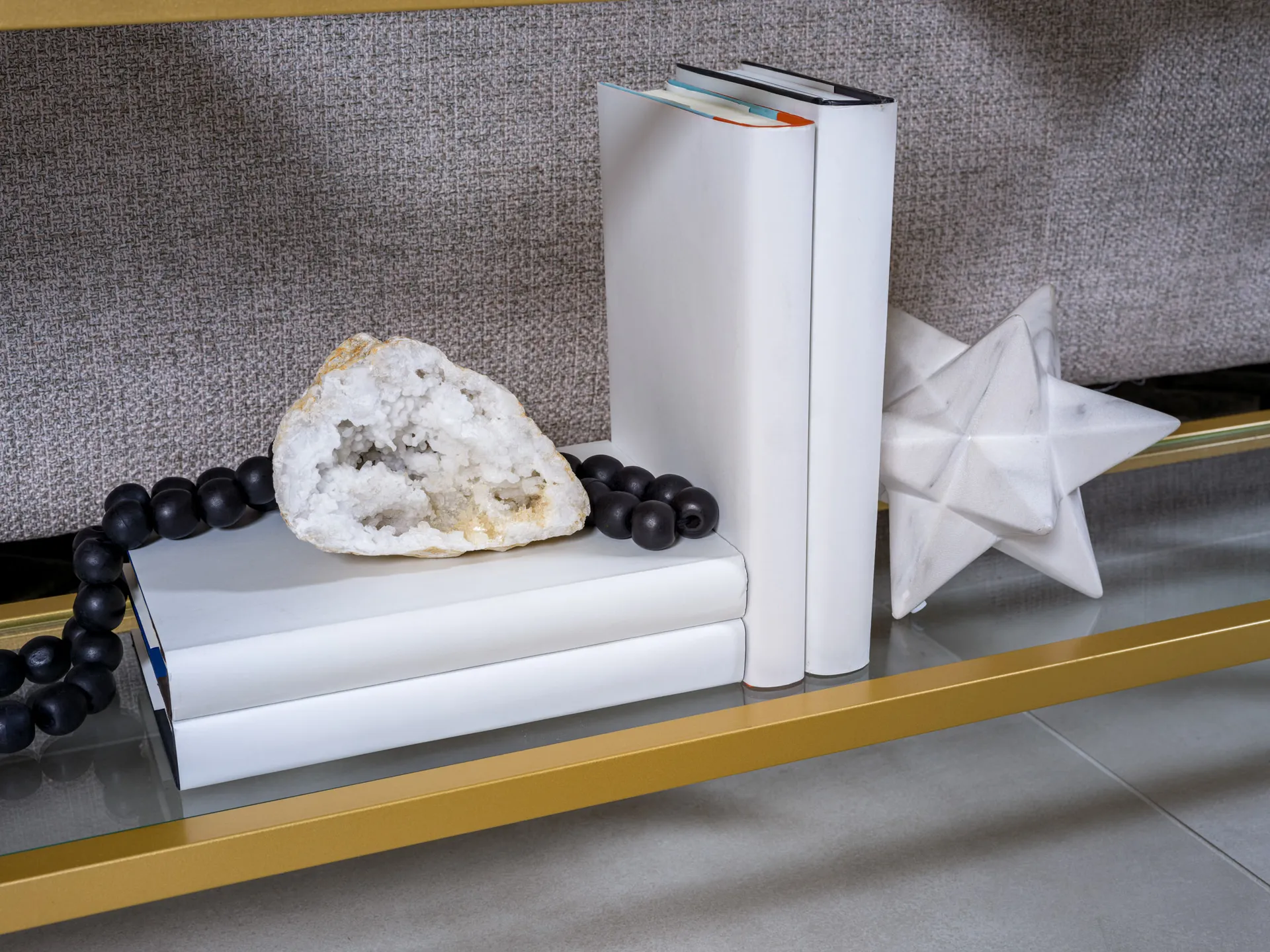 A gold-framed shelf with white books, a crystal geode, black beads, and a white star sculpture for decoration.