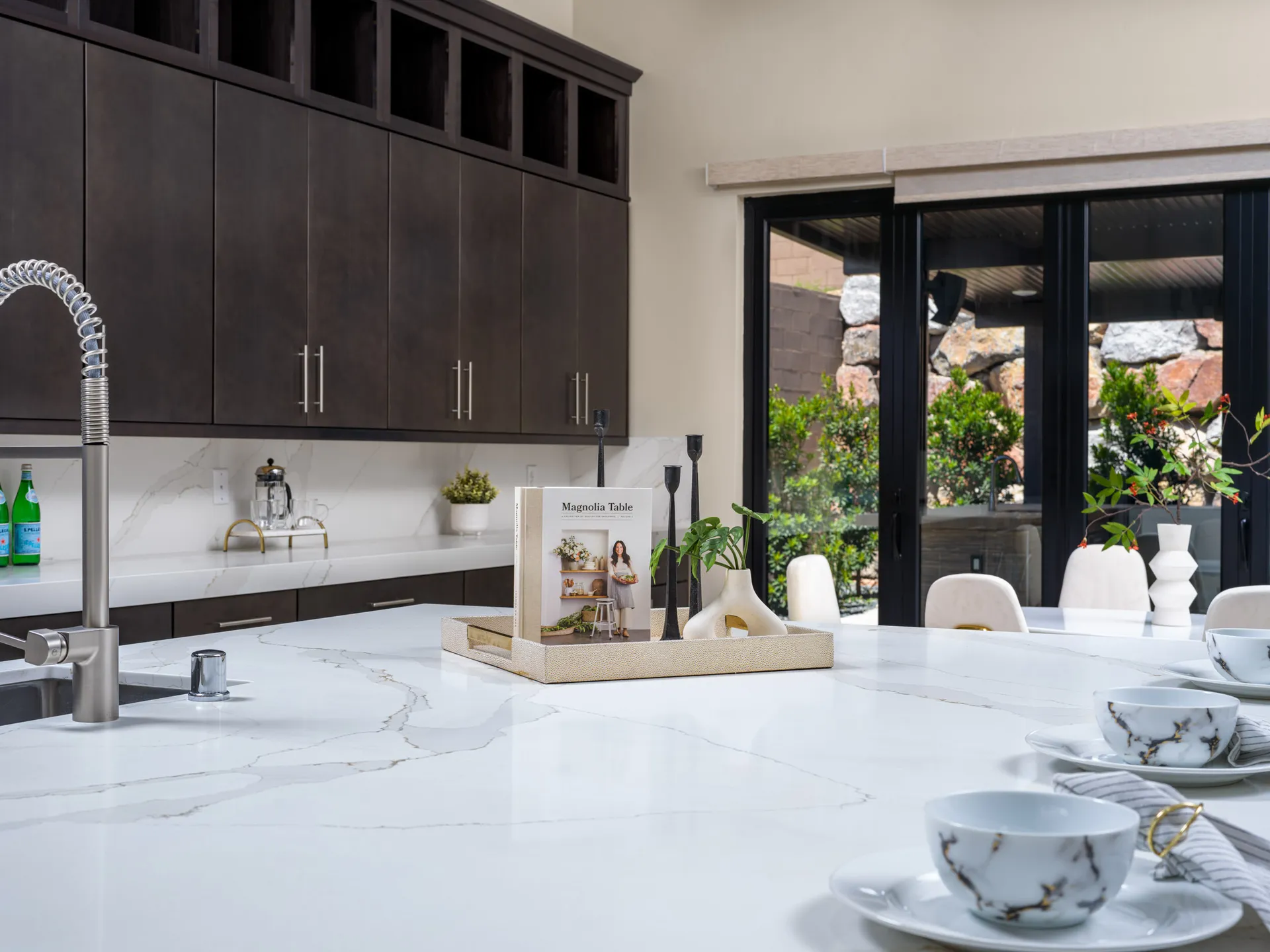 A spacious kitchen with a large white island, a decorative tray holding a cookbook and vases, and sliding doors leading to an outdoor patio.