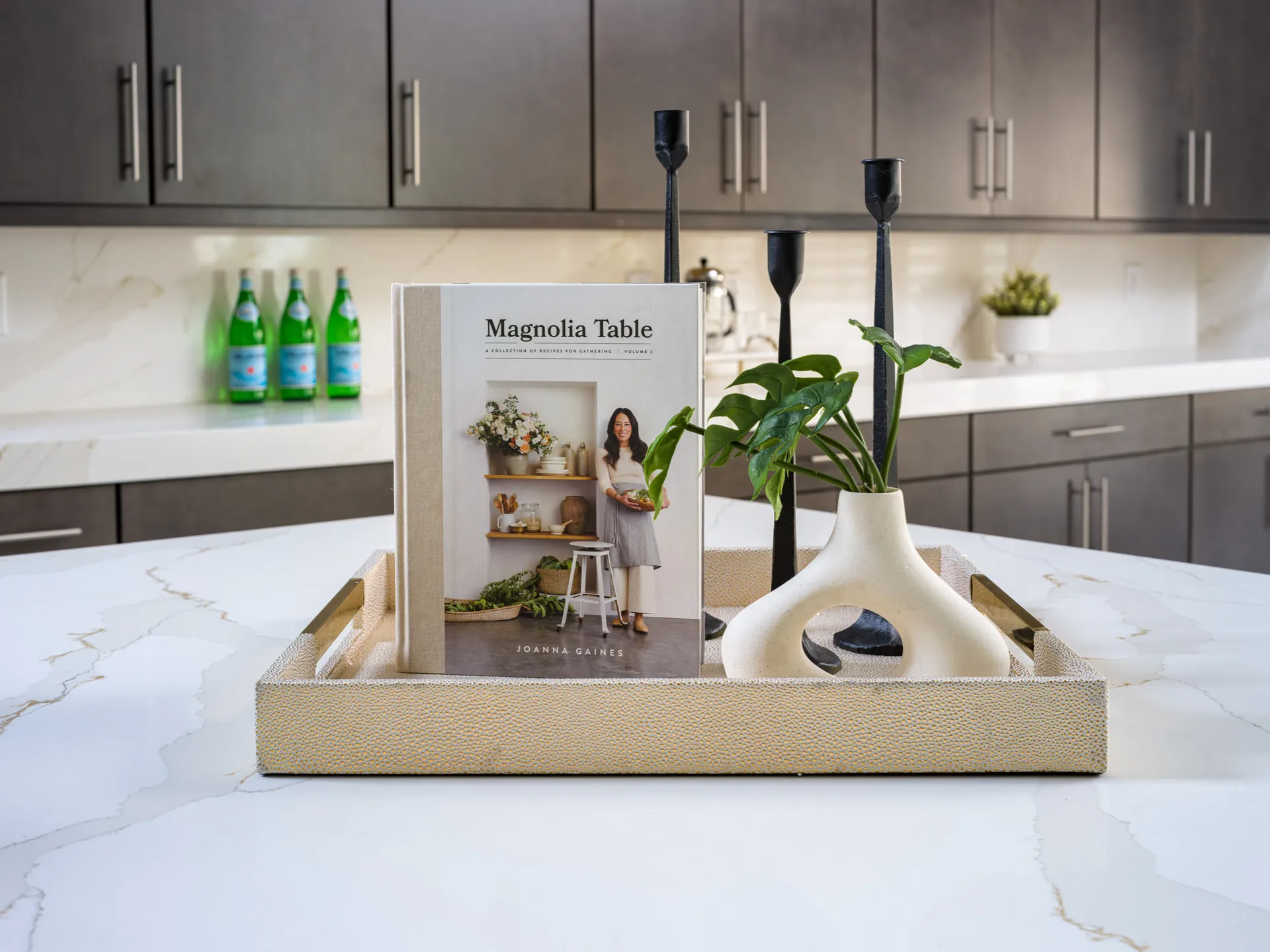 A neutral tray on a white kitchen island featuring a cookbook, black candle holders, and a modern vase with green leaves.