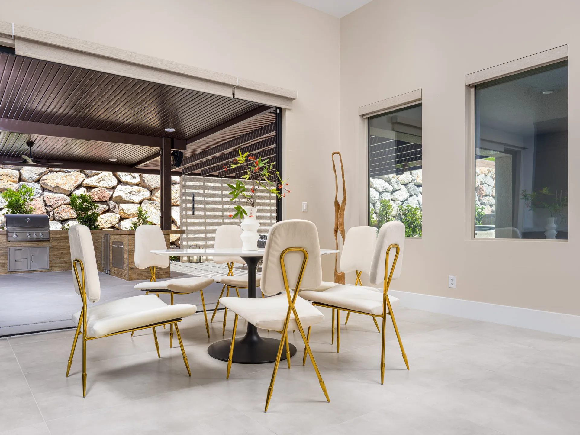 A bright dining area with white chairs featuring gold frames around a round table, opening to an outdoor patio with stone walls and a built-in grill.