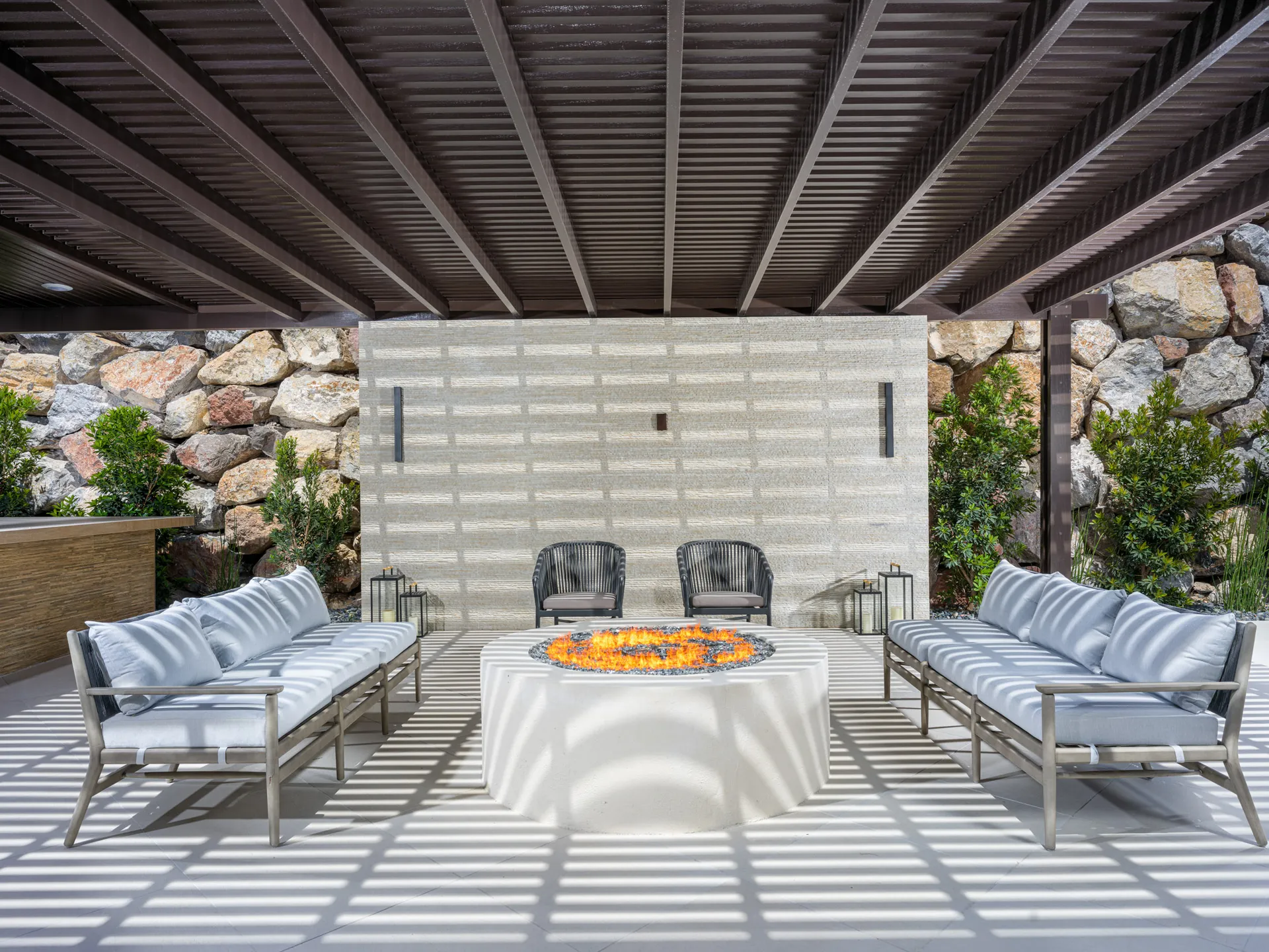 An outdoor seating area under a pergola with gray sofas around a lit fire pit, set against a stacked stone wall and lush greenery.