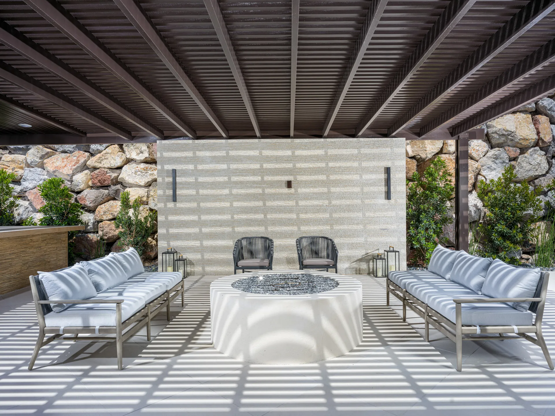 A covered patio with sleek gray seating arranged around a circular fire pit. Stone walls and greenery complete the inviting outdoor setting.