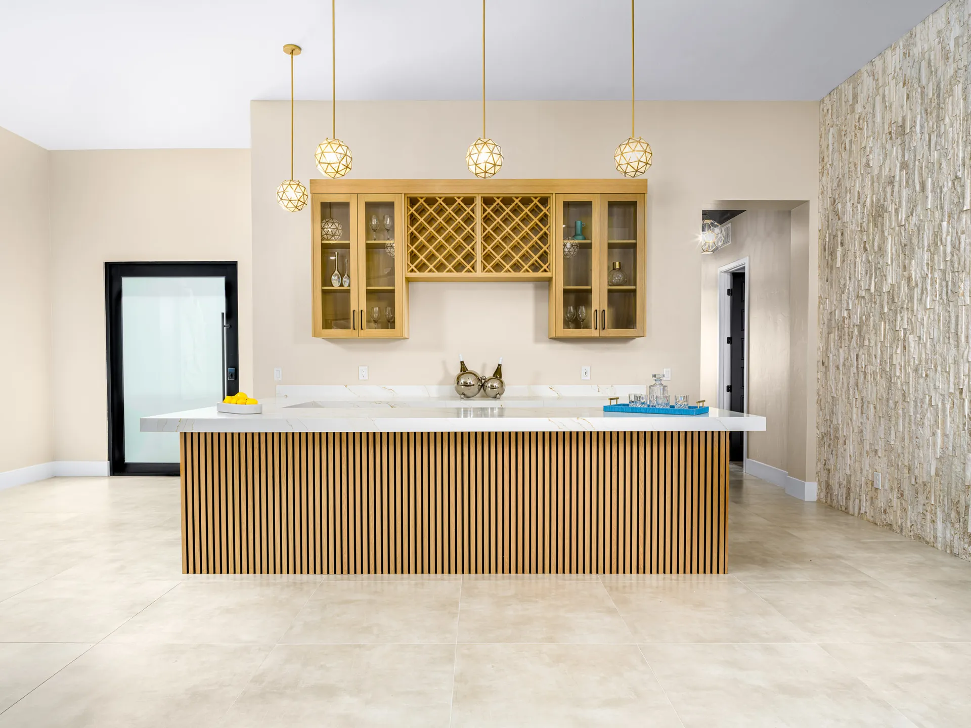 A contemporary bar with a slatted wood panel front, gold pendant lights, and a stone accent wall - staged by Stately in Las Vegas for refined entertaining.