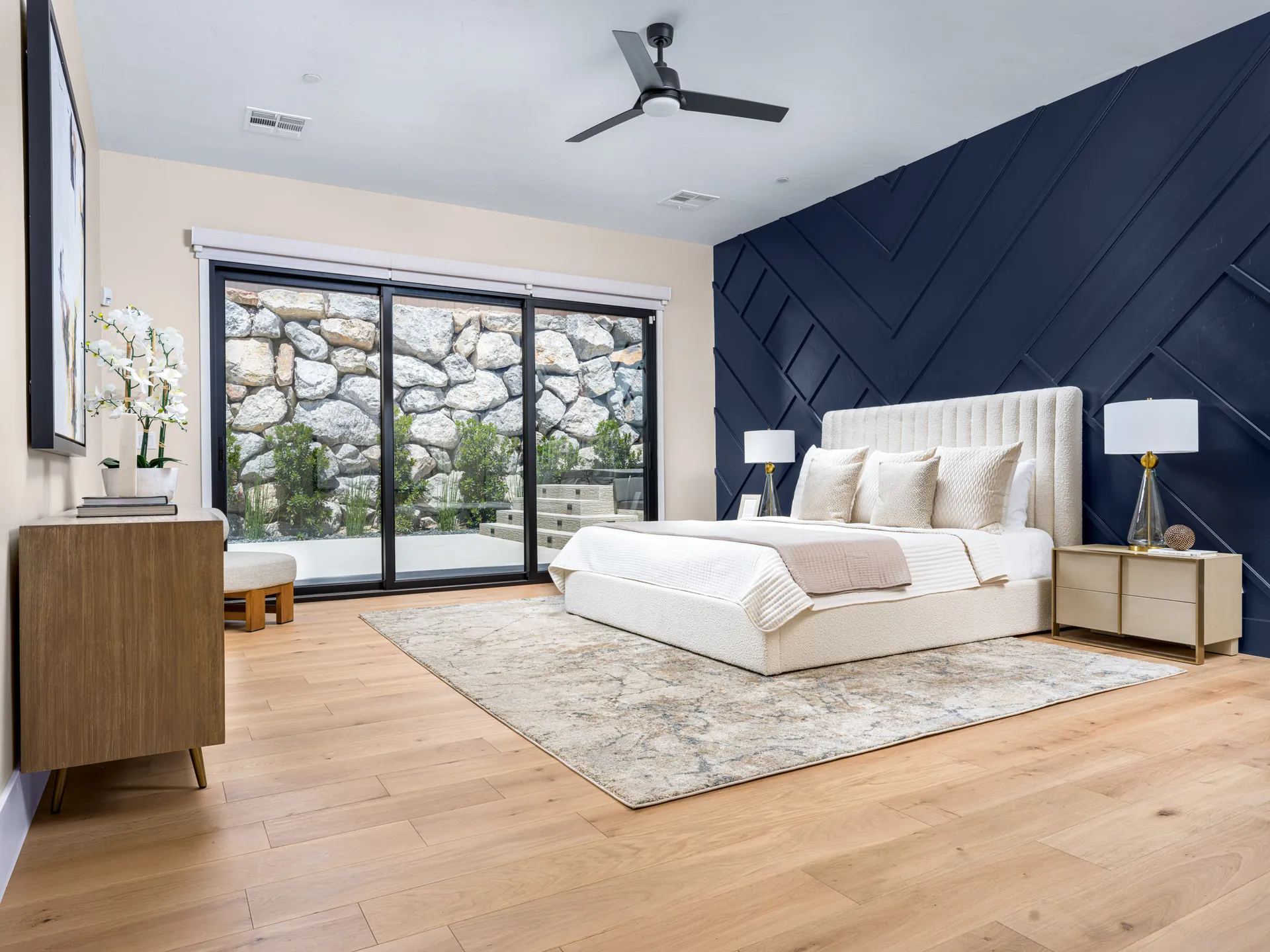 A cream upholstered bed with neutral bedding against a navy geometric accent wall, large glass doors opening to a stone-lined courtyard - staged by Stately in Las Vegas.