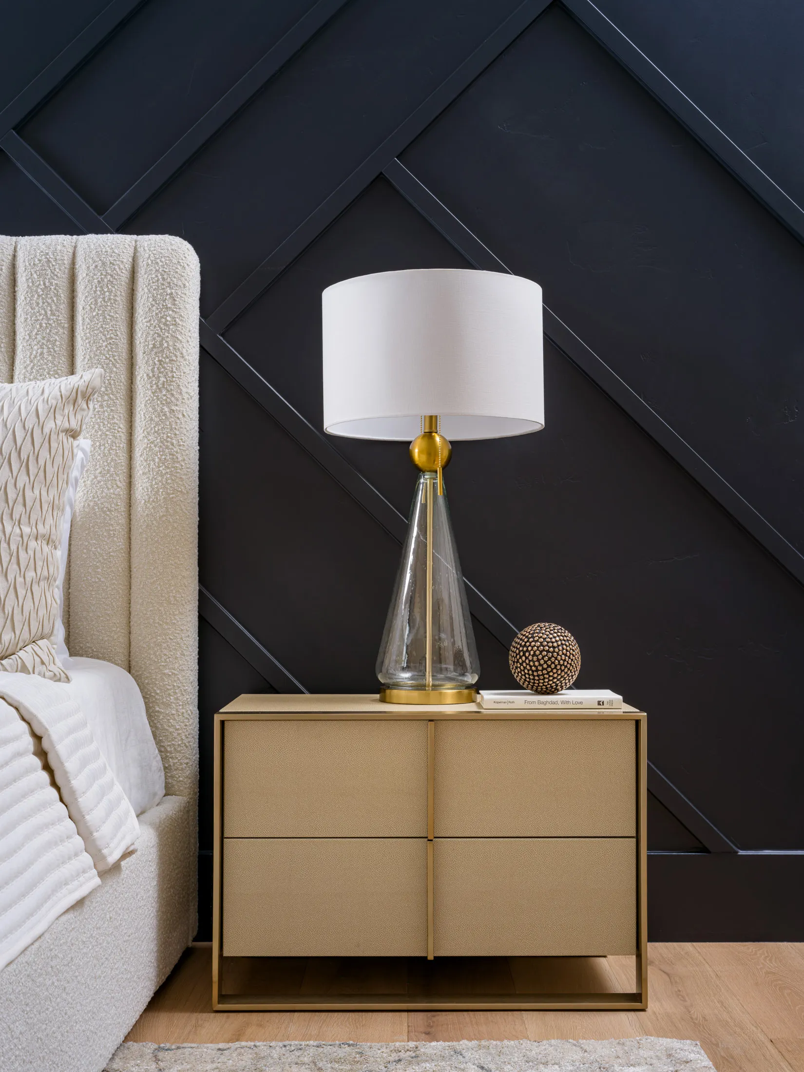 A plush cream bed with a gold nightstand and glass lamp, set against a dark geometric accent wall, styled by Stately for a sleek bedroom design