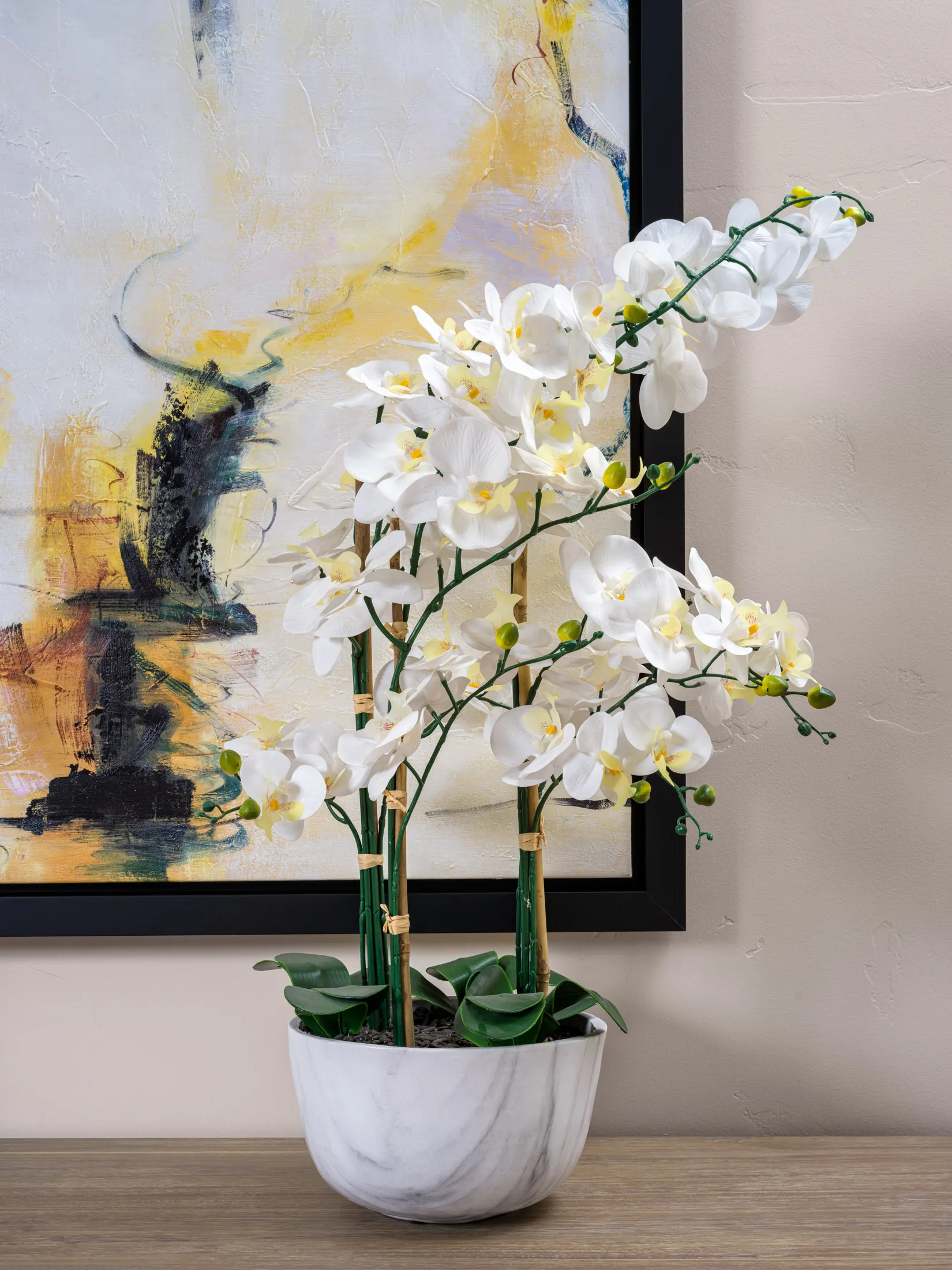 A white orchid arrangement in a marble pot on a wooden console, set against an abstract painting with warm yellow tones. Staged by Stately in Las Vegas for a refined interior accent.