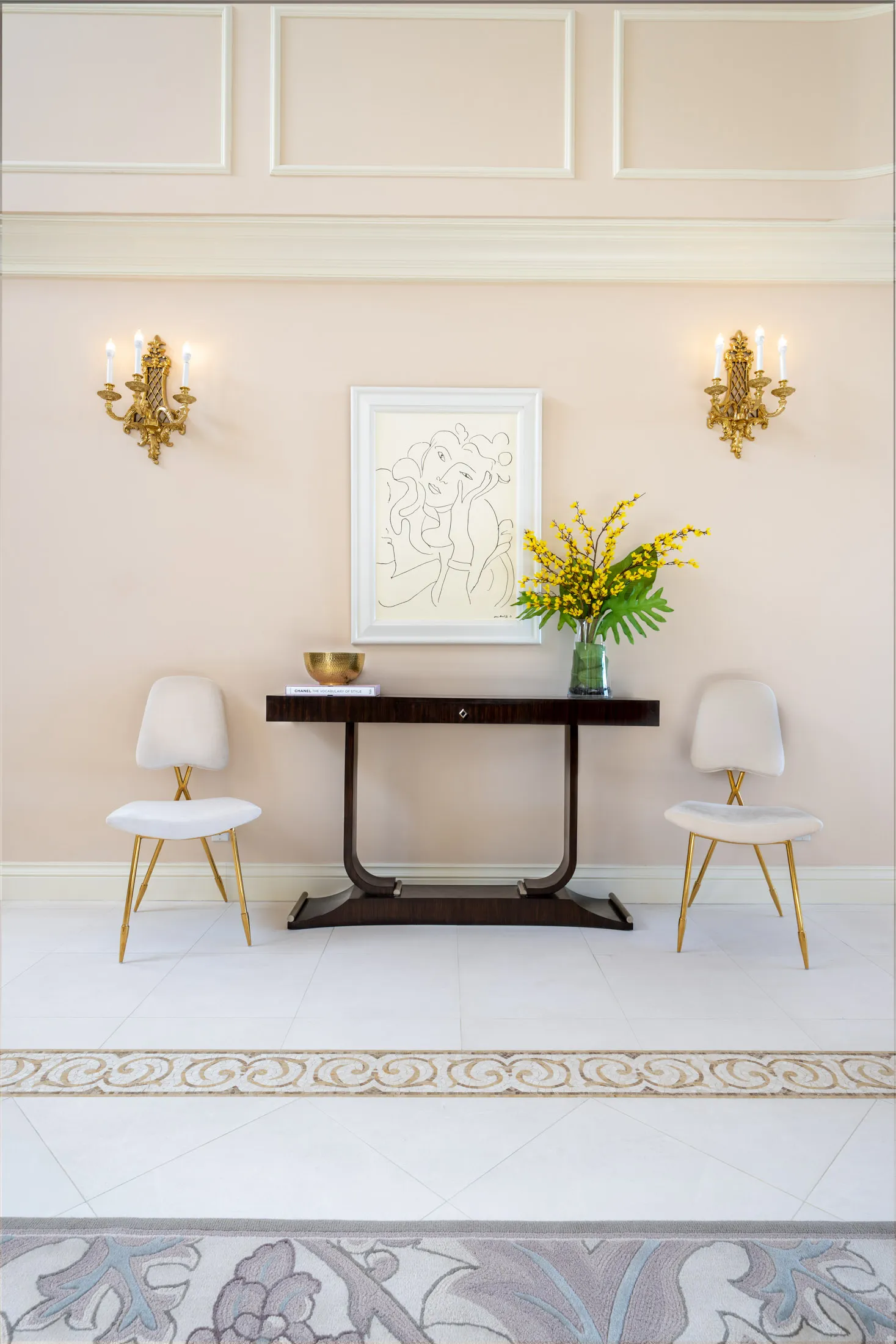 A dark wood console table centered beneath a line-art painting, flanked by two white chairs with gold legs. Ornate wall sconces and a vibrant floral arrangement accent the neutral setting.