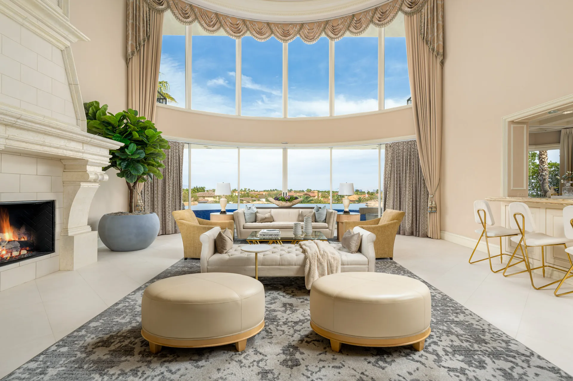A spacious living room with a curved wall of tall windows, a grand stone fireplace on one side, and plush neutral seating. Large potted greenery and round ottomans complete the open layout.