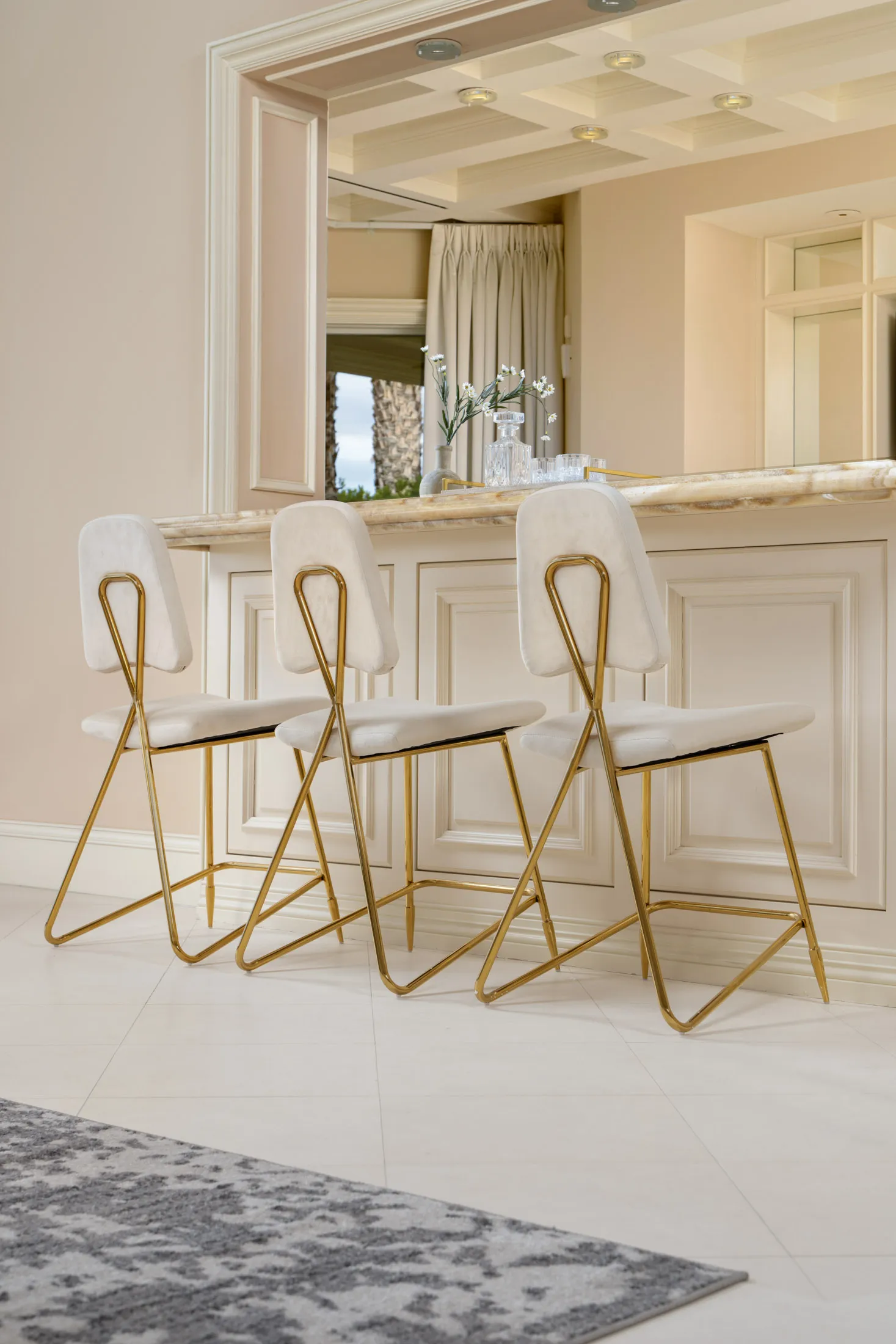 Three gold-framed bar stools with white velvet seats lined up at a light-colored counter. The design features a marble-like countertop and neutral cabinetry.