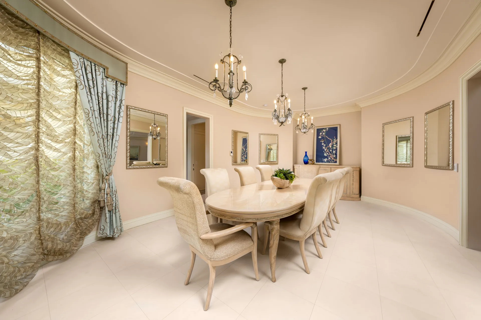 A neutral dining room with an oval table and upholstered chairs beneath ornate chandeliers. Flowing drapes and mirrored accents lend a luxurious feel.