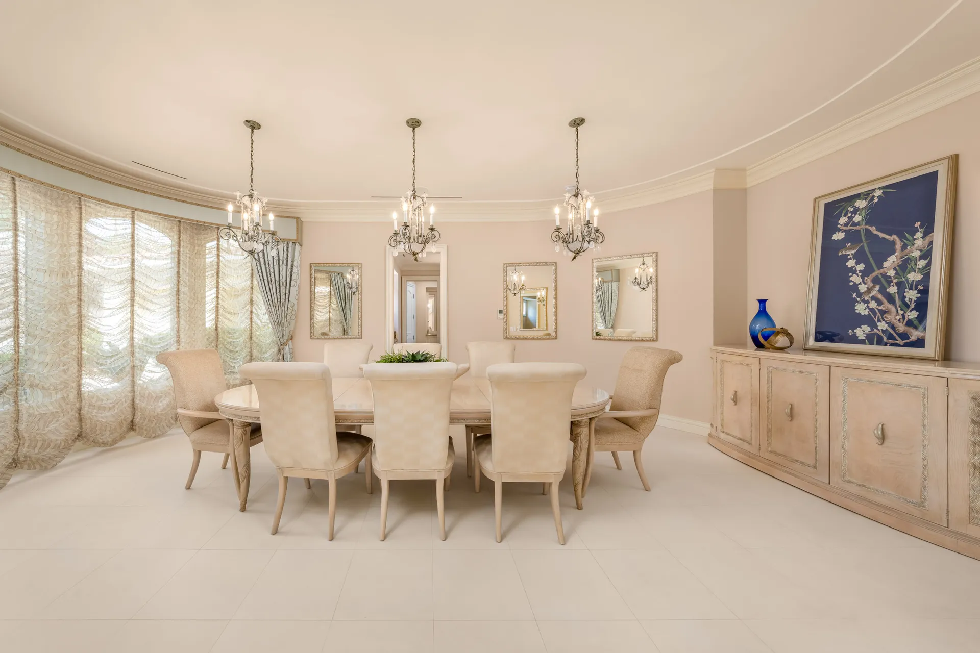 A round dining table surrounded by upholstered chairs under two ornate chandeliers. Curved drapes and mirrors add a soft, elegant backdrop.