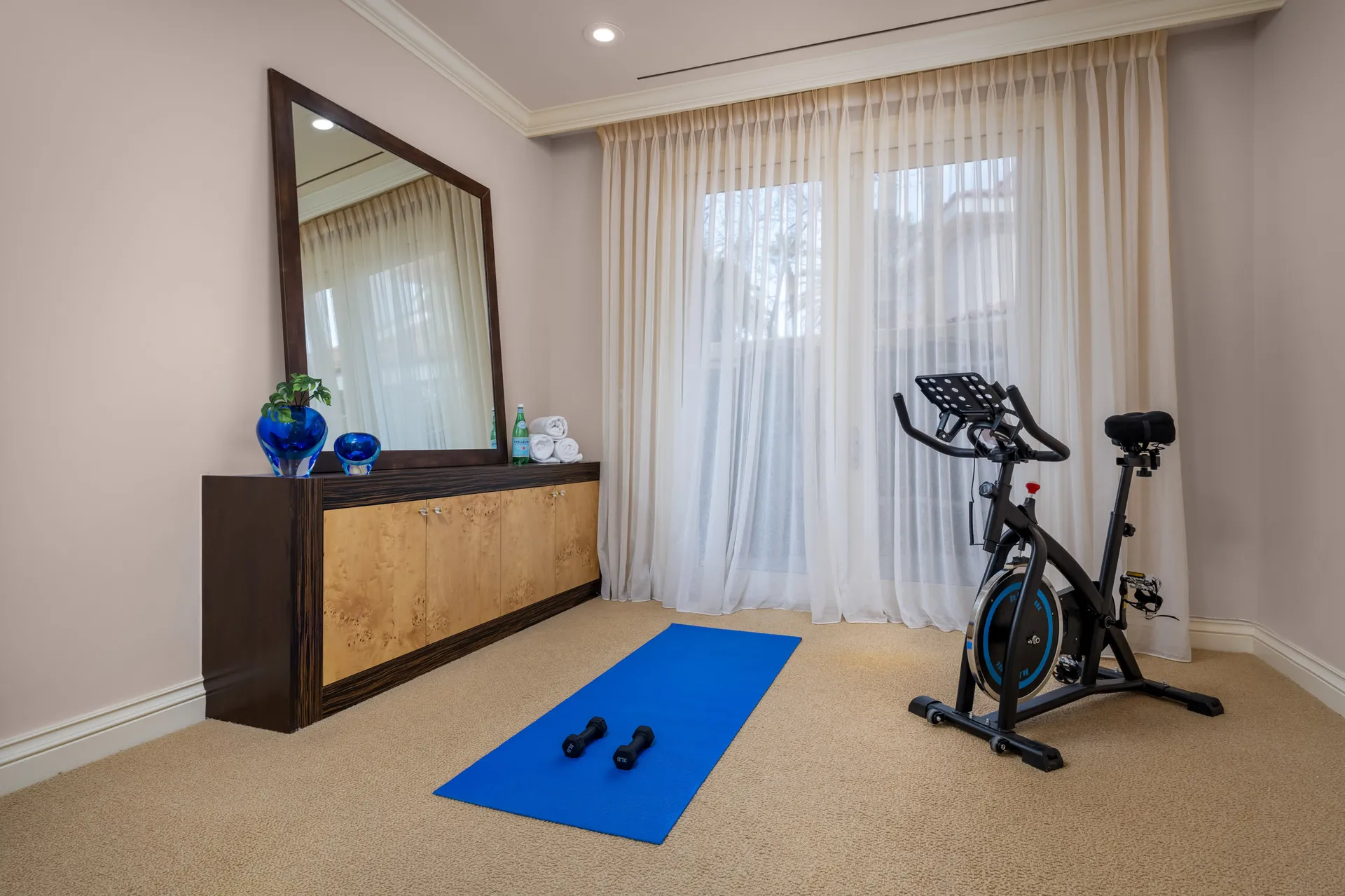 A compact workout space featuring a stationary bike, a blue yoga mat with weights, and a large mirror above a wooden console. Sheer curtains filter natural light.